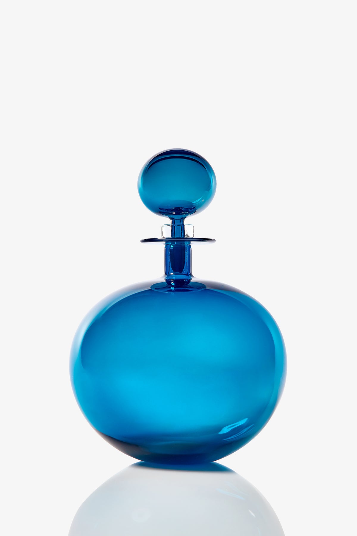 The Small Angelic Bottle, a round blue glass perfume bottle with a matching spherical stopper, sits on a reflective white surface against a plain background.