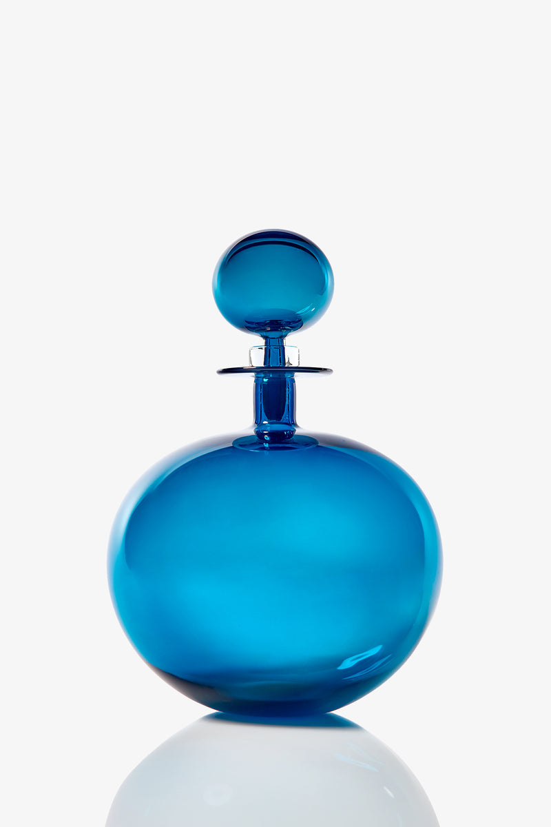 The Small Angelic Bottle, a round blue glass perfume bottle with a matching spherical stopper, sits on a reflective white surface against a plain background.
