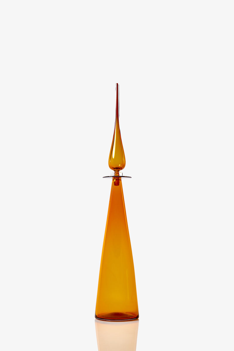 The Petite Straight Cone Decanter is a tall amber glass bottle with a geometric triangular base and slender pointed stopper, shown against a plain white background.