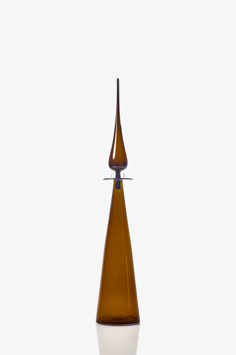 The Petite Straight Cone Decanter is a tall, slender brown glass bottle with a long, pointed stopper and a modern, minimalist design. Its reflective surface stands out against the plain white background.