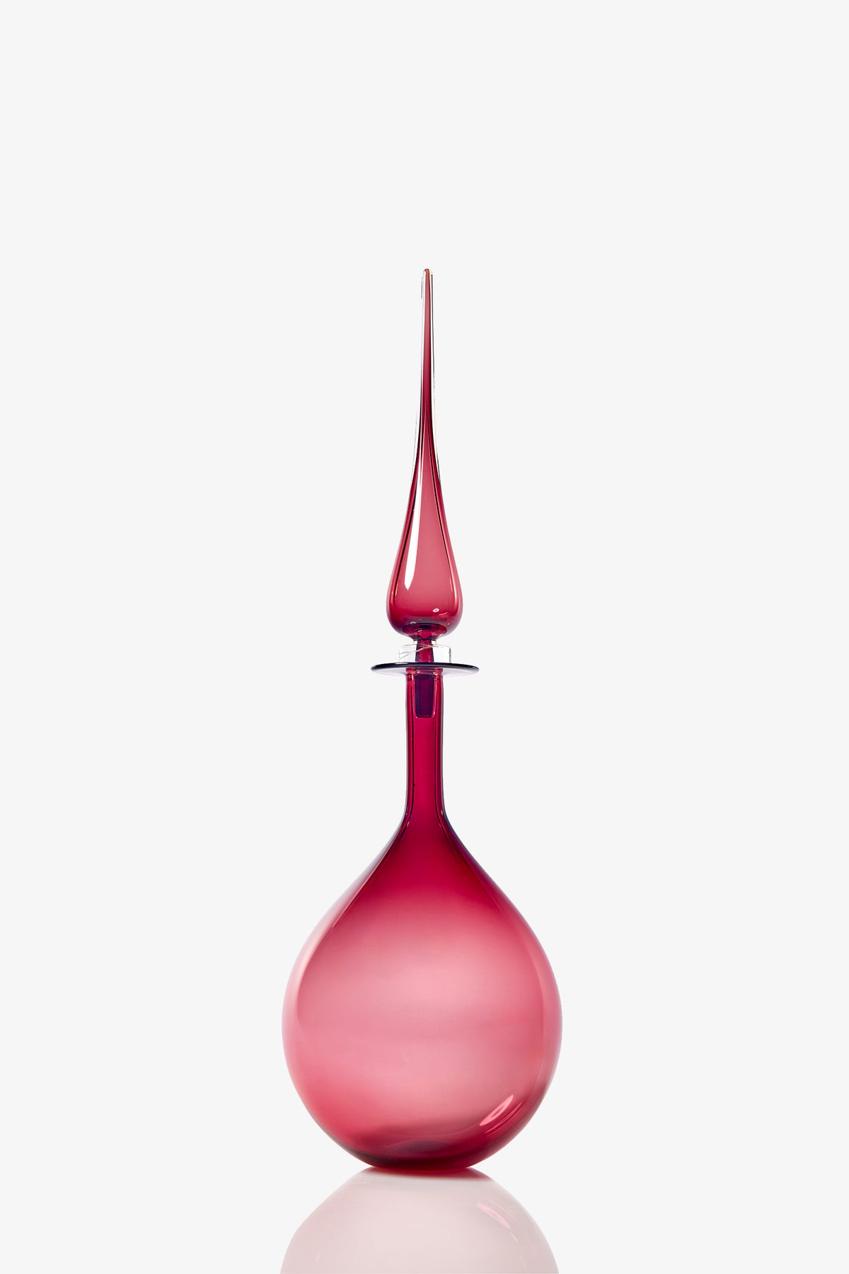 The Petite Tear Drop Decanter is a tall, elegant vessel with a round base and a long, pointed stopper, crafted from translucent pink glass and shown against a plain white background.