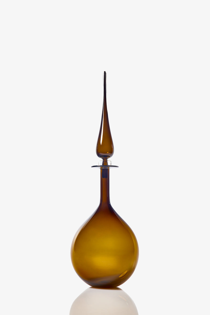 The Petite Tear Drop Decanter, a brown glass bottle with a round base and tall, slender pointed stopper, is photographed against a white background with a subtle reflection below.