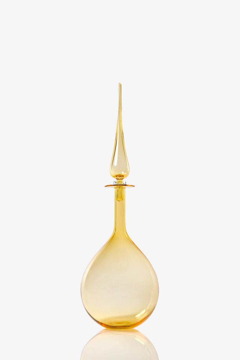 The Petite Tear Drop Decanter, made from transparent golden yellow glass, features a tall teardrop-shaped body and a long pointed stopper, displayed against a plain white background.