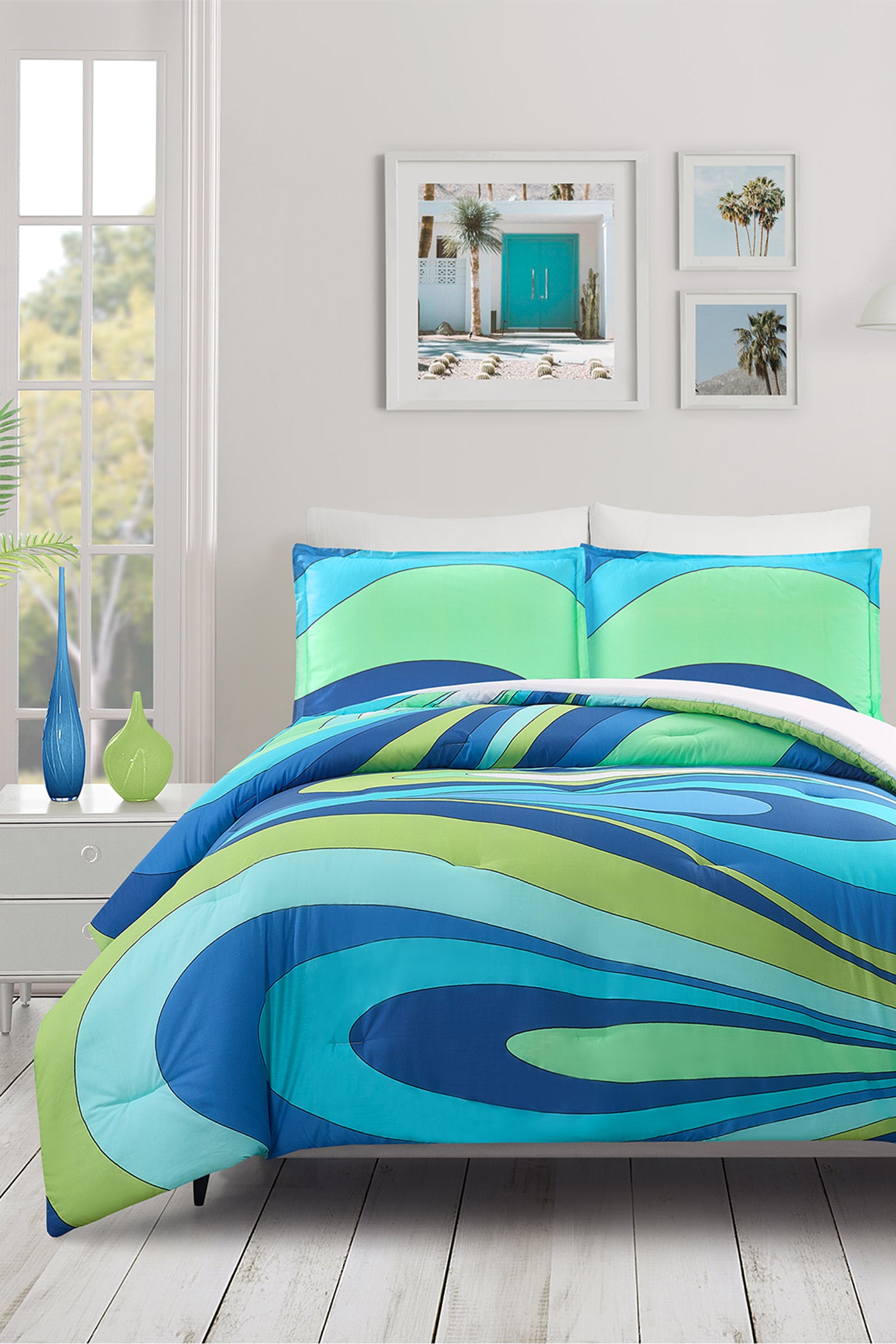 A modern bedroom features a blue Wynwood Waves King 3-Piece Comforter Set, white walls, a large window, a white nightstand with blue and green vases, and framed beach-themed artwork above the bed.