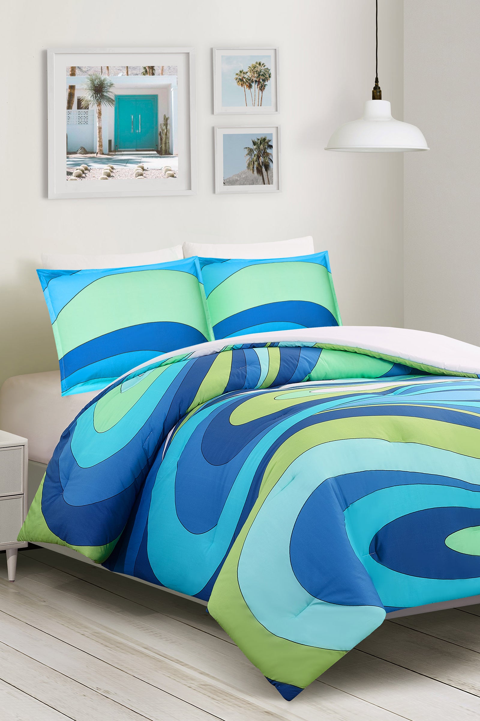 A modern white bedroom features the Wynwood Waves King 3-Piece Comforter Set in a blue and green wavy pattern with matching pillows, palm tree photos on the wall, and a white pendant light above the headboard.