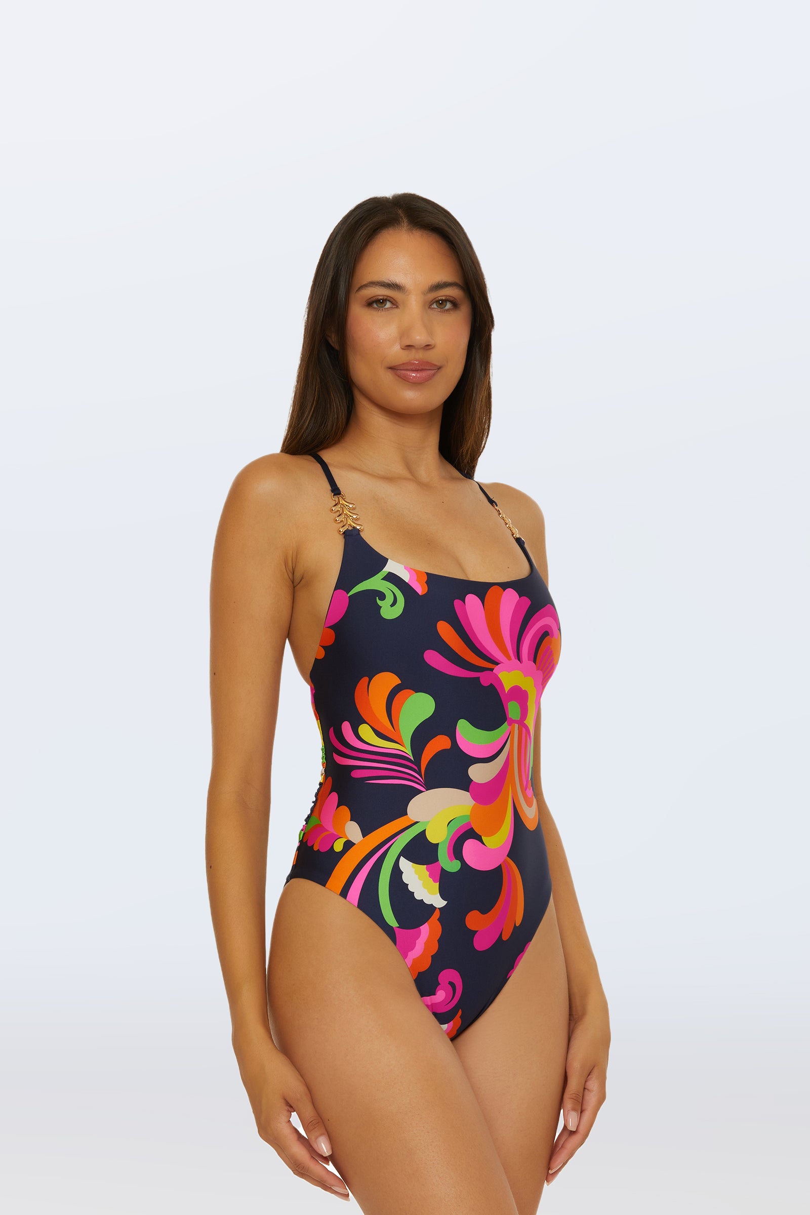 A woman models the Twist Back One-Piece Swimsuit - Palms Paradise, featuring vibrant multicolored abstract floral patterns on dark blue, standing against a plain light background and smiling slightly at the camera.