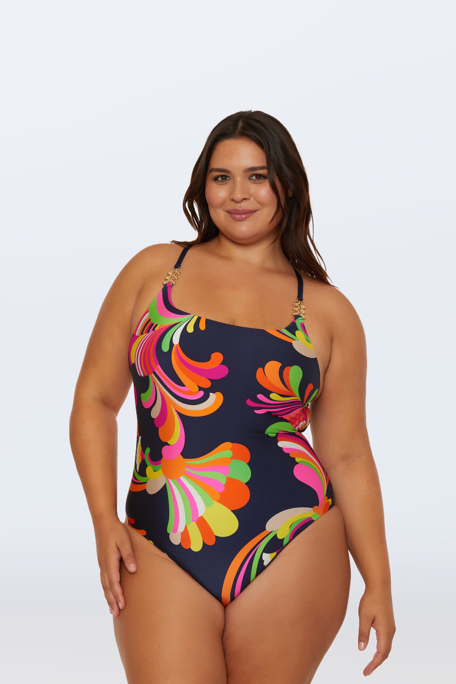 A woman in the Twist Back One-Piece Swimsuit - Palms Paradise, featuring a colorful abstract pattern and spaghetti straps, stands against a plain light background, smiling with her arms relaxed at her sides.