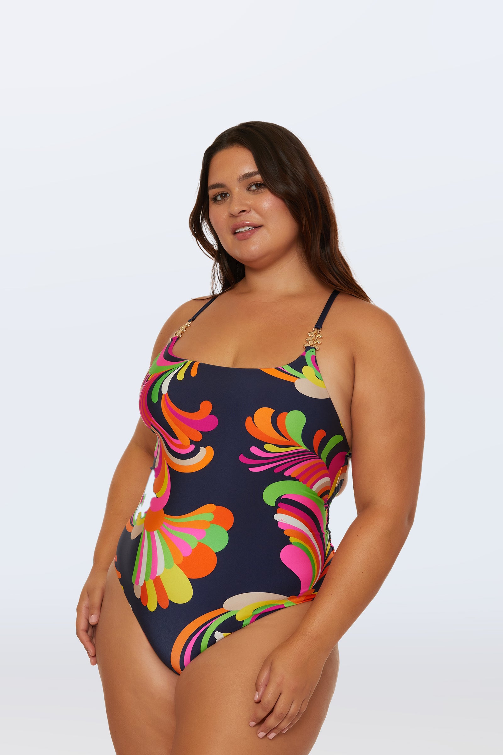 A woman with long brown hair wears the Twist Back One-Piece Swimsuit - Palms Paradise, a colorful, retro-patterned suit with thin straps, standing confidently against a plain light background.