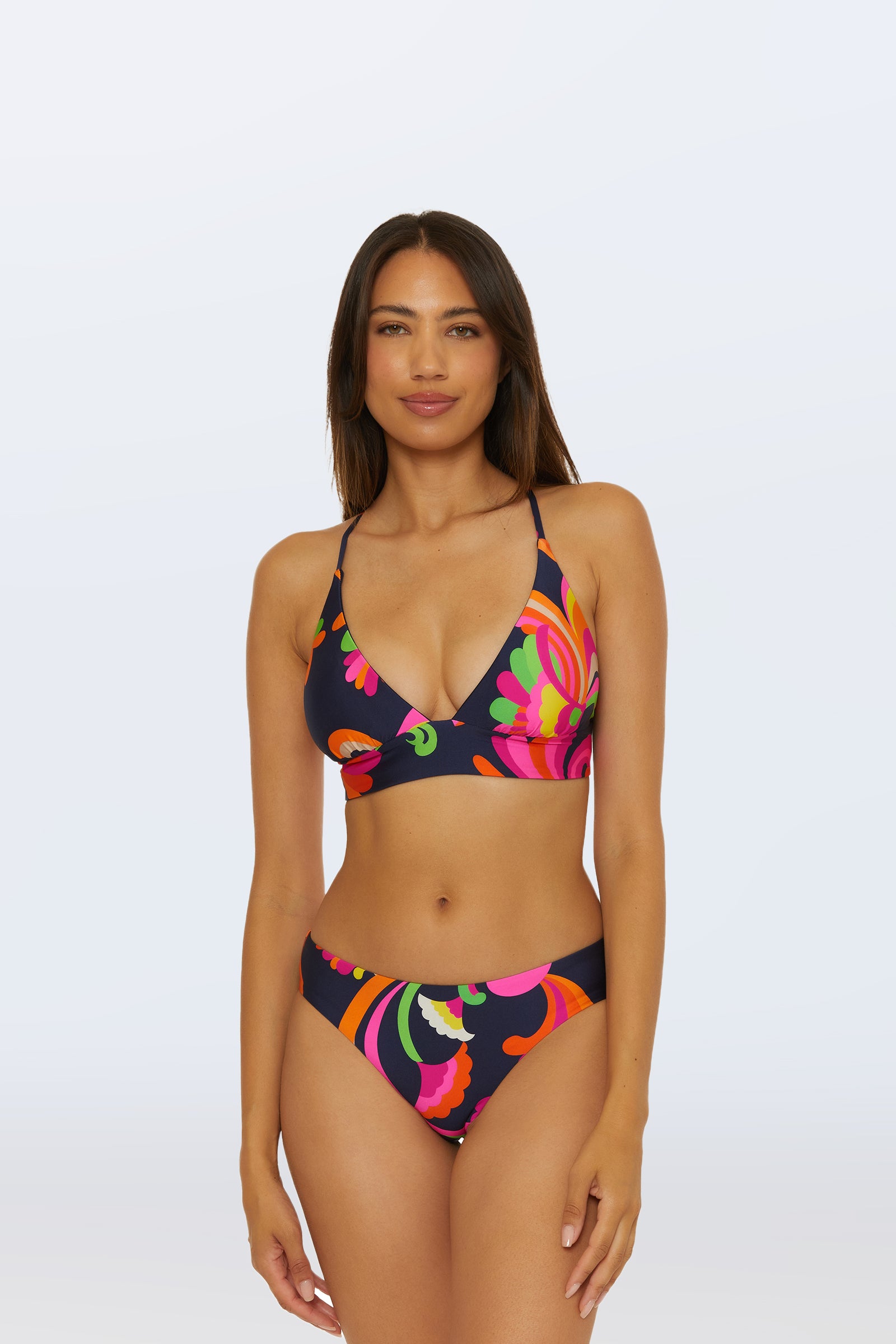 A woman wears the Reversible Hipster Bottom - Palms Paradise bikini, featuring bold pink, orange, green, and yellow floral and swirl patterns, standing against a light background.