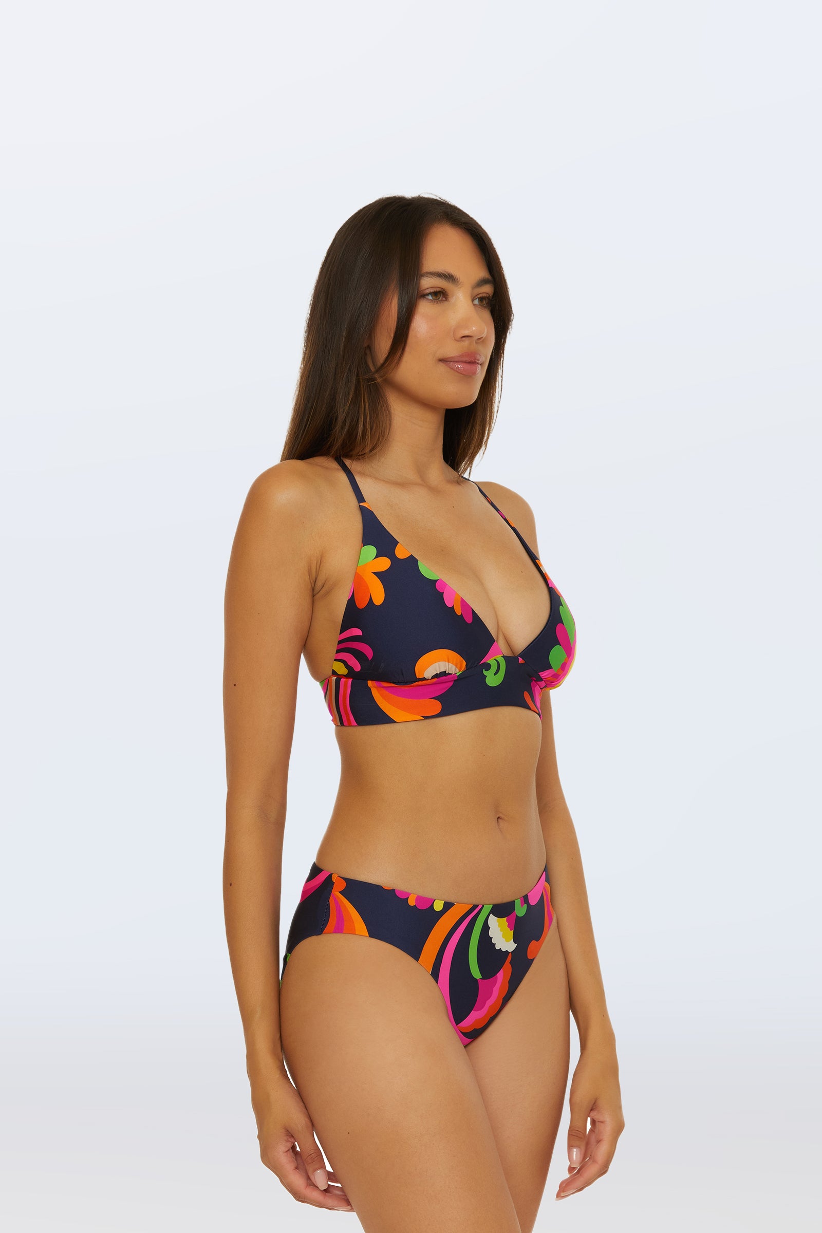 A woman with long, straight brown hair stands against a plain background wearing the Reversible Hipster Bottom - Palms Paradise, a dark bikini bottom featuring a colorful retro floral pattern in pink, orange, and green. She looks slightly to the side.