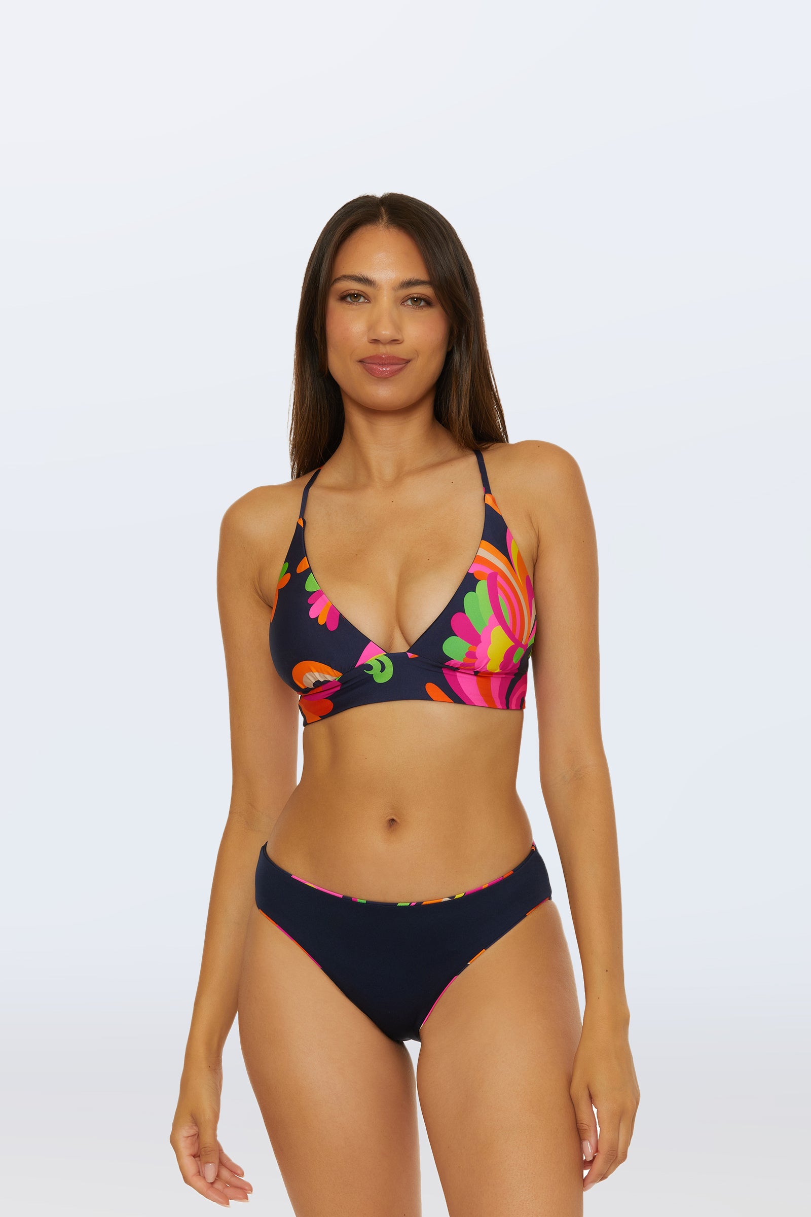A woman with long brown hair wears the Reversible Hipster Bottom - Palms Paradise bikini, featuring a navy blue color and colorful floral print, standing against a plain light background.