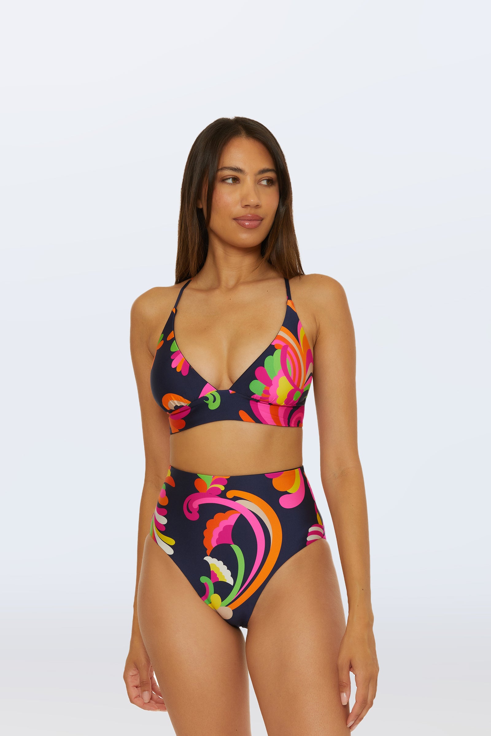 A woman in the Reversible High Waist Bottom - Palms Paradise bikini, featuring a bold floral and swirl print, stands against a plain light background with long straight hair and a neutral expression, gazing slightly to the side.