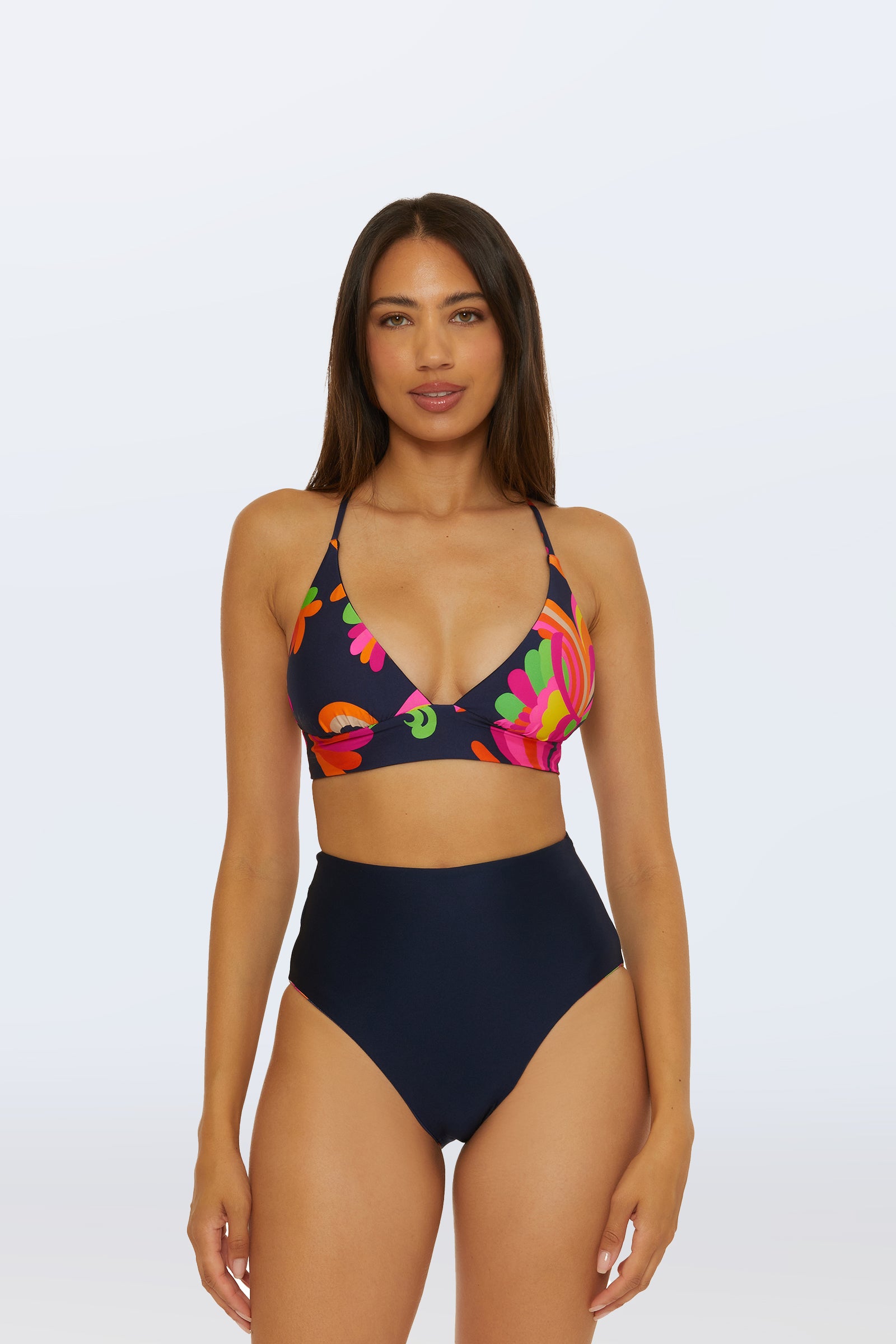 A woman stands facing forward, wearing the Reversible High Waist Bottom - Palms Paradise in navy blue, paired with a navy blue bikini top featuring colorful floral patterns on a light background.