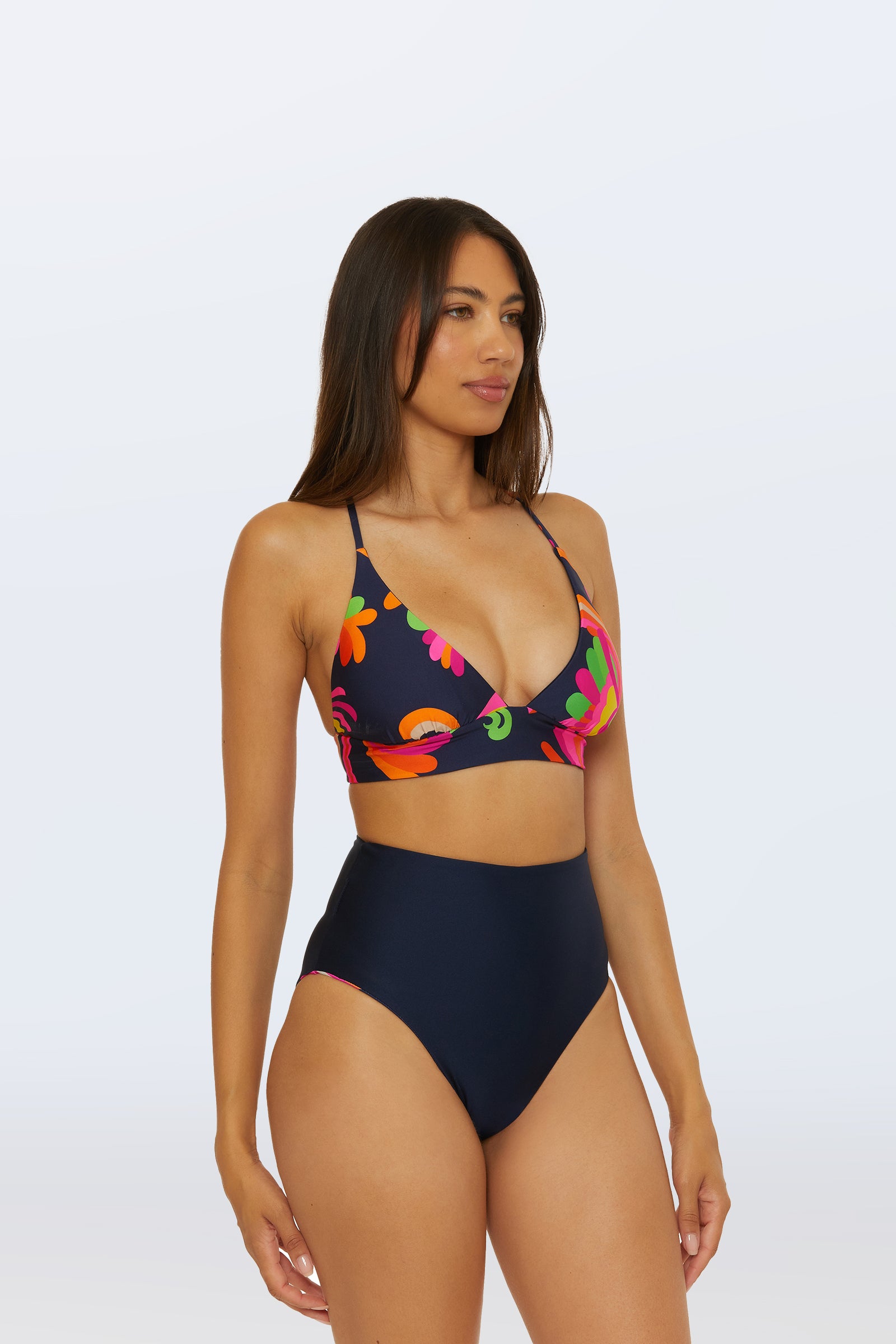 A woman with long brown hair wears a dark floral bikini top and the Reversible High Waist Bottom - Palms Paradise, standing against a plain light background.