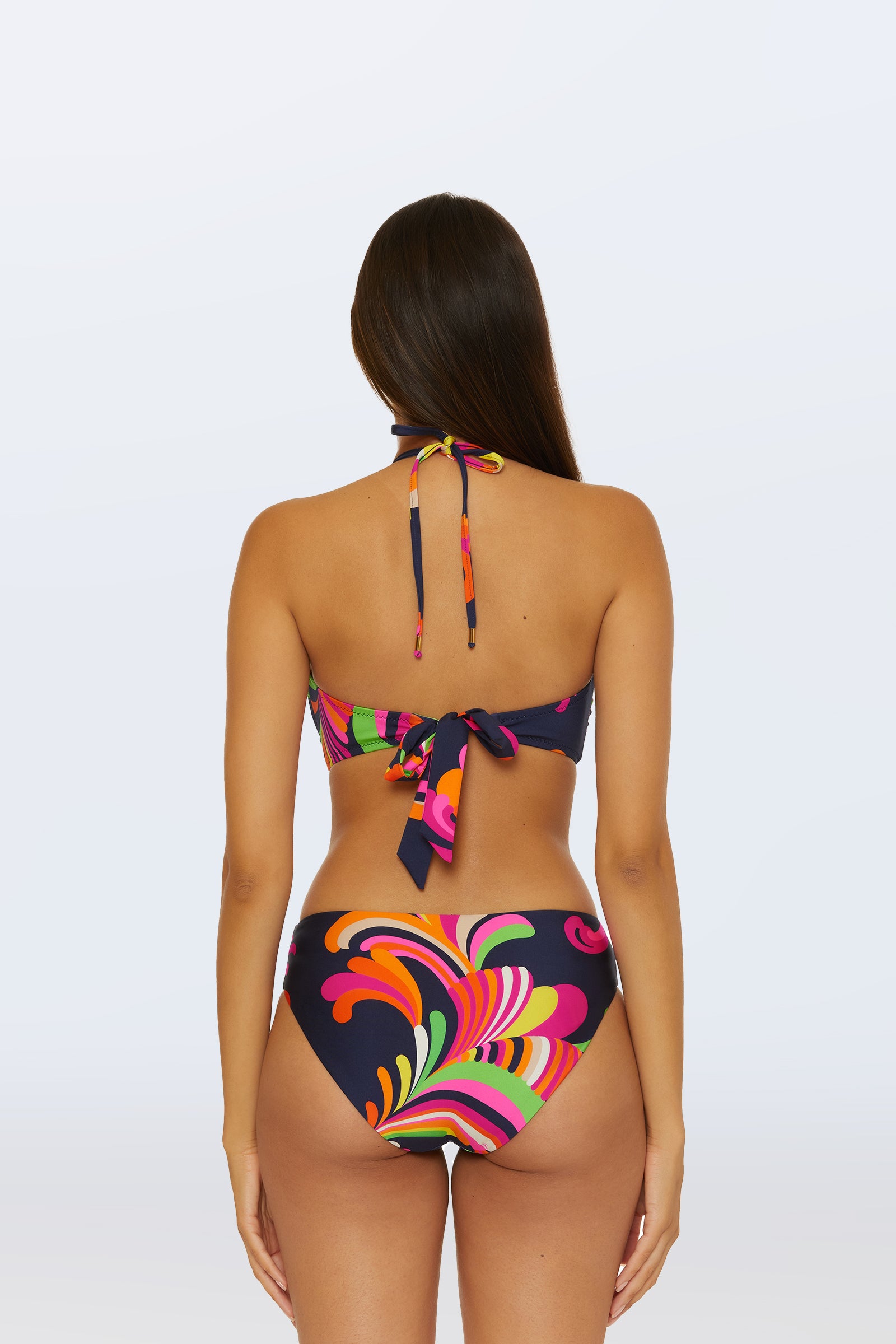 A woman with long brown hair models the Bandeau Swim Top - Palms Paradise, featuring a vibrant abstract print in pink, green, orange, and yellow on a dark background. The bikini ties at the back and neck; she is shown from behind.