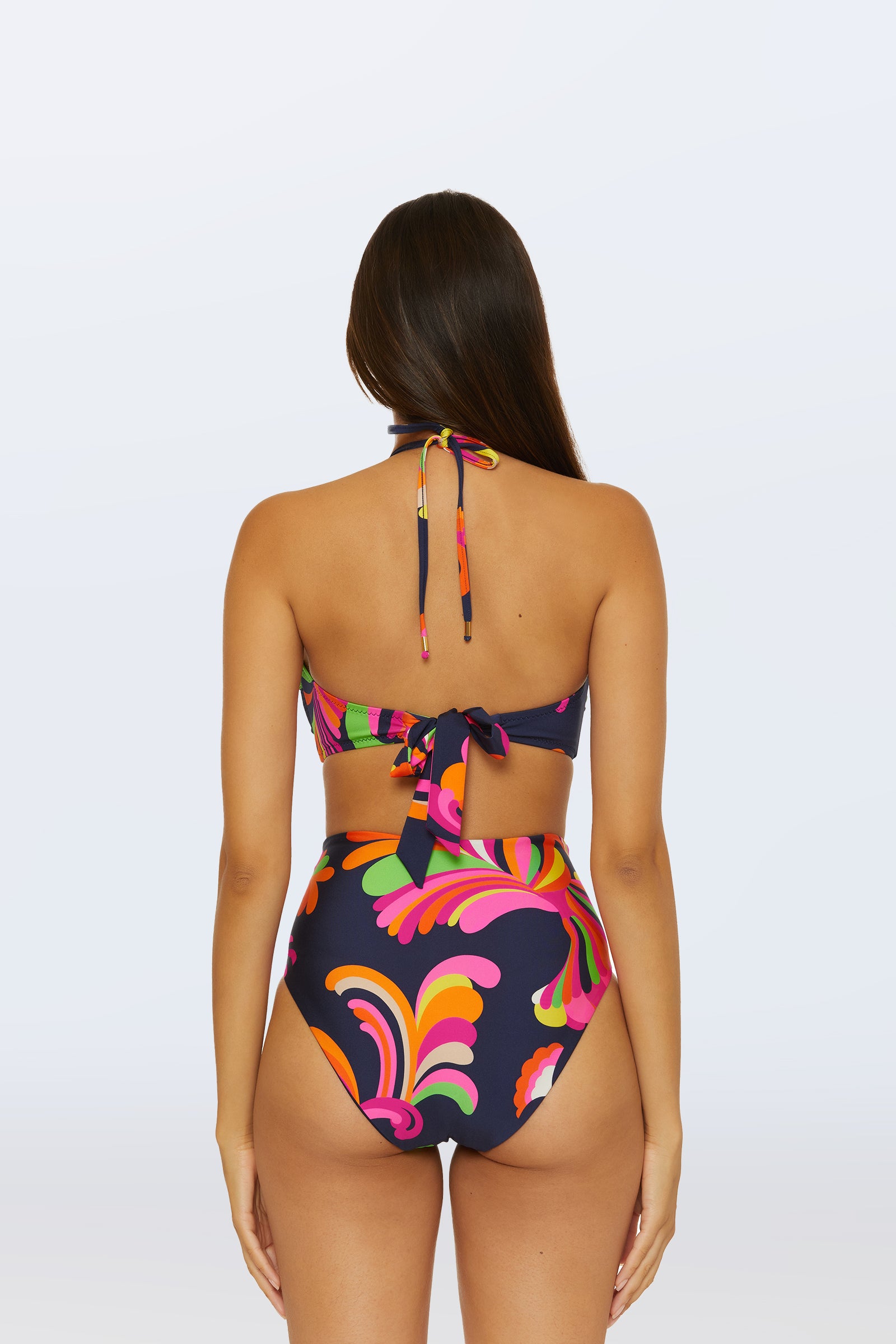 A woman with long brown hair is seen from the back wearing the Palms Paradise Bandeau Swim Top, featuring a colorful retro pattern and a halter-neck tie, against a plain light background.