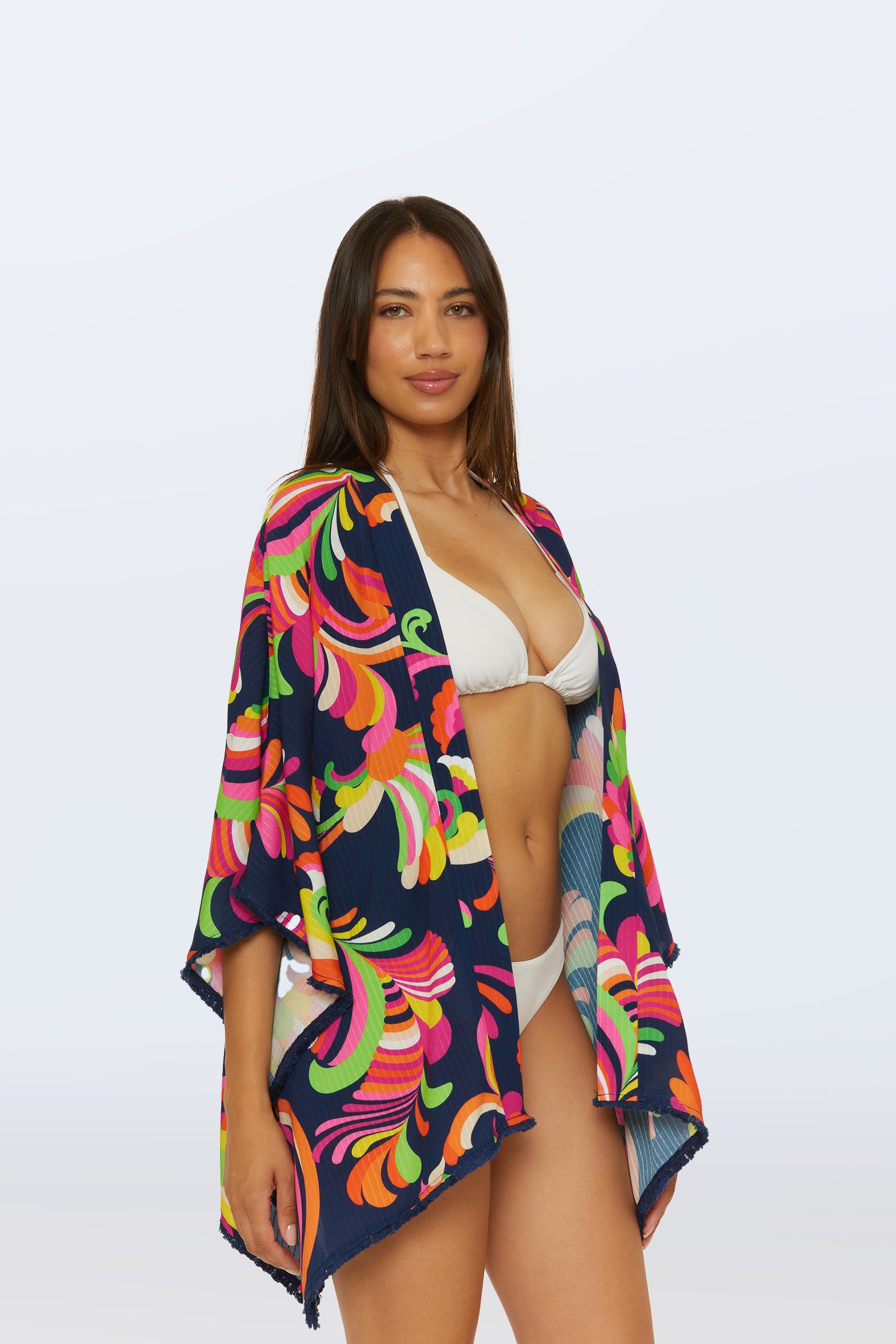 A woman wears a white bikini and the Kimono Swim Cover-Up - Palms Paradise, featuring vibrant floral and swirl patterns in navy blue, standing confidently against a plain, light background.