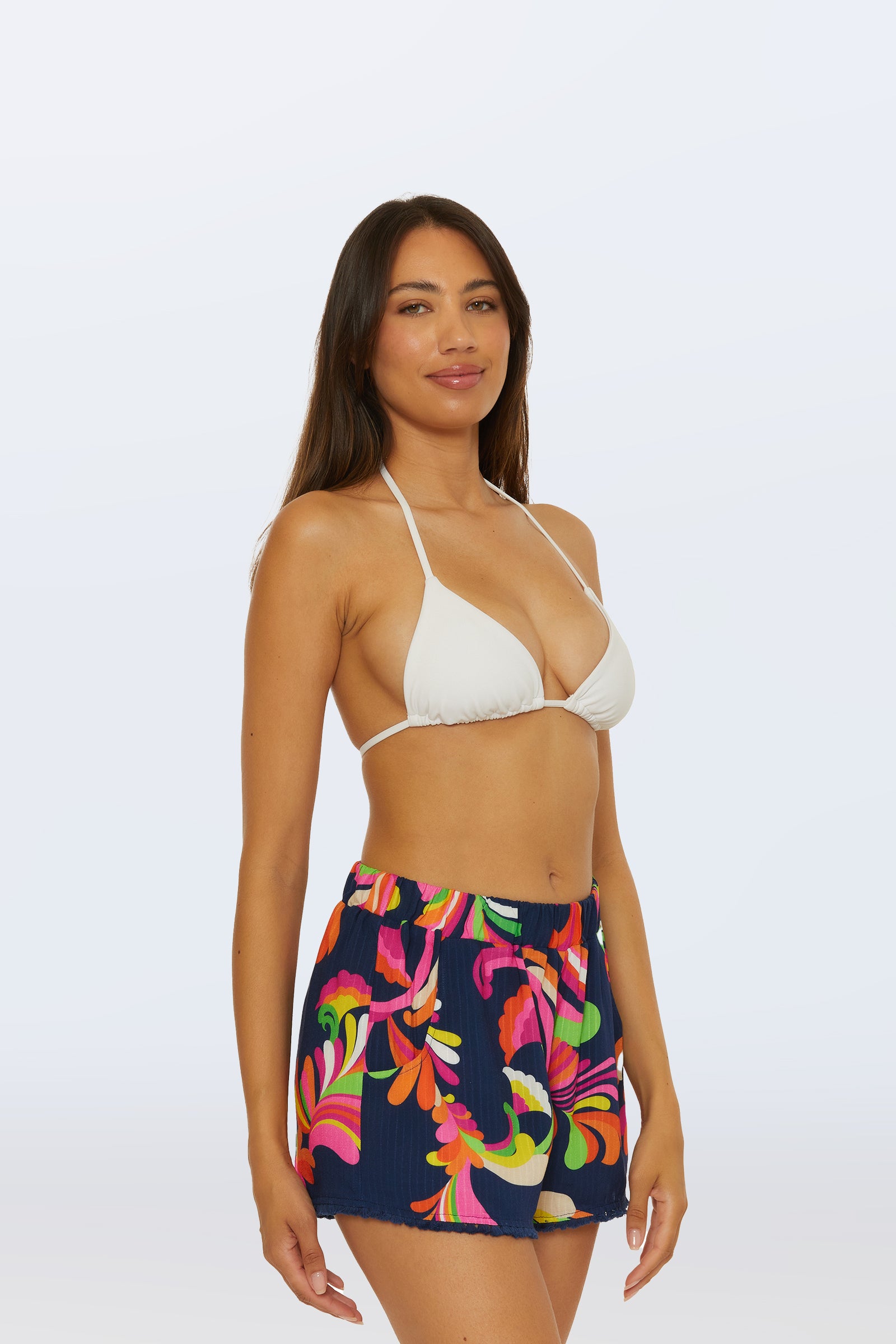 A woman smiles gently with her arms relaxed at her sides, wearing the Cover-Up Short - Palms Paradise—a navy blue short with a colorful floral pattern—and a white bikini top, standing against a plain light background.