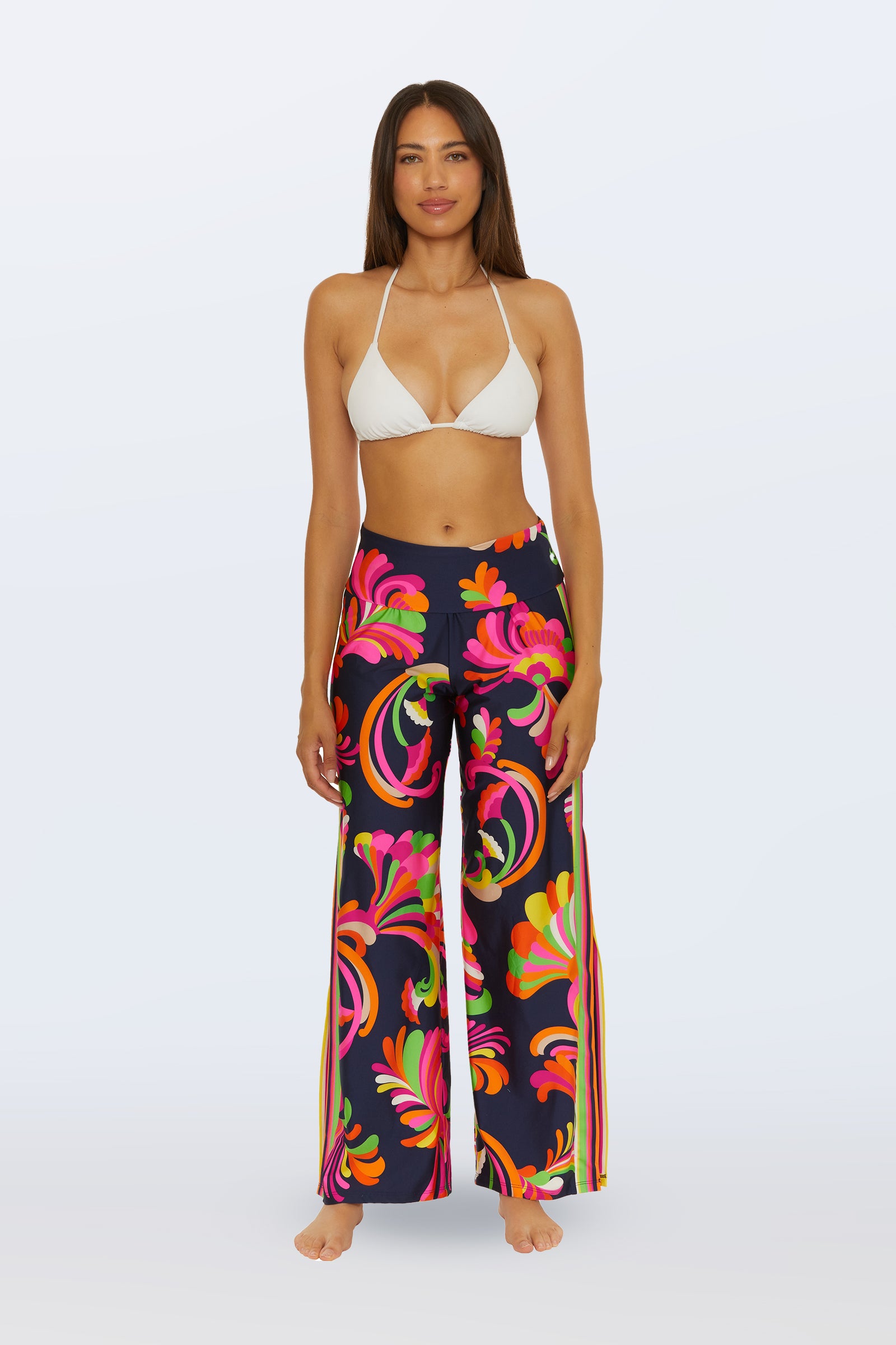 A woman stands barefoot facing forward against a plain light backdrop, wearing a white bikini top and the Swim Cover-Up Pant - Palms Paradise in Multi, featuring high-waisted, wide-leg design with a bold floral pattern.