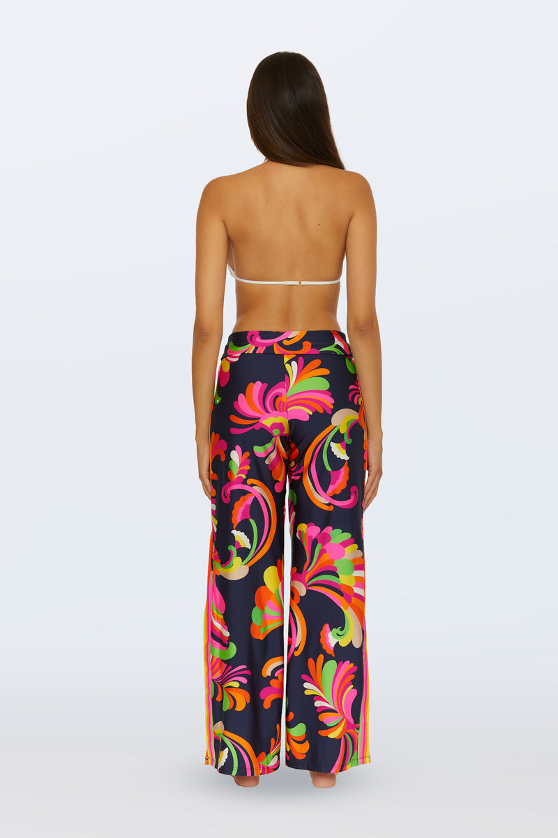 A woman stands barefoot facing away, wearing the Swim Cover-Up Pant - Palms Paradise, featuring a vibrant abstract palm print and wide-leg, high-waisted design. She pairs it with a white halter bikini top against a plain light background.