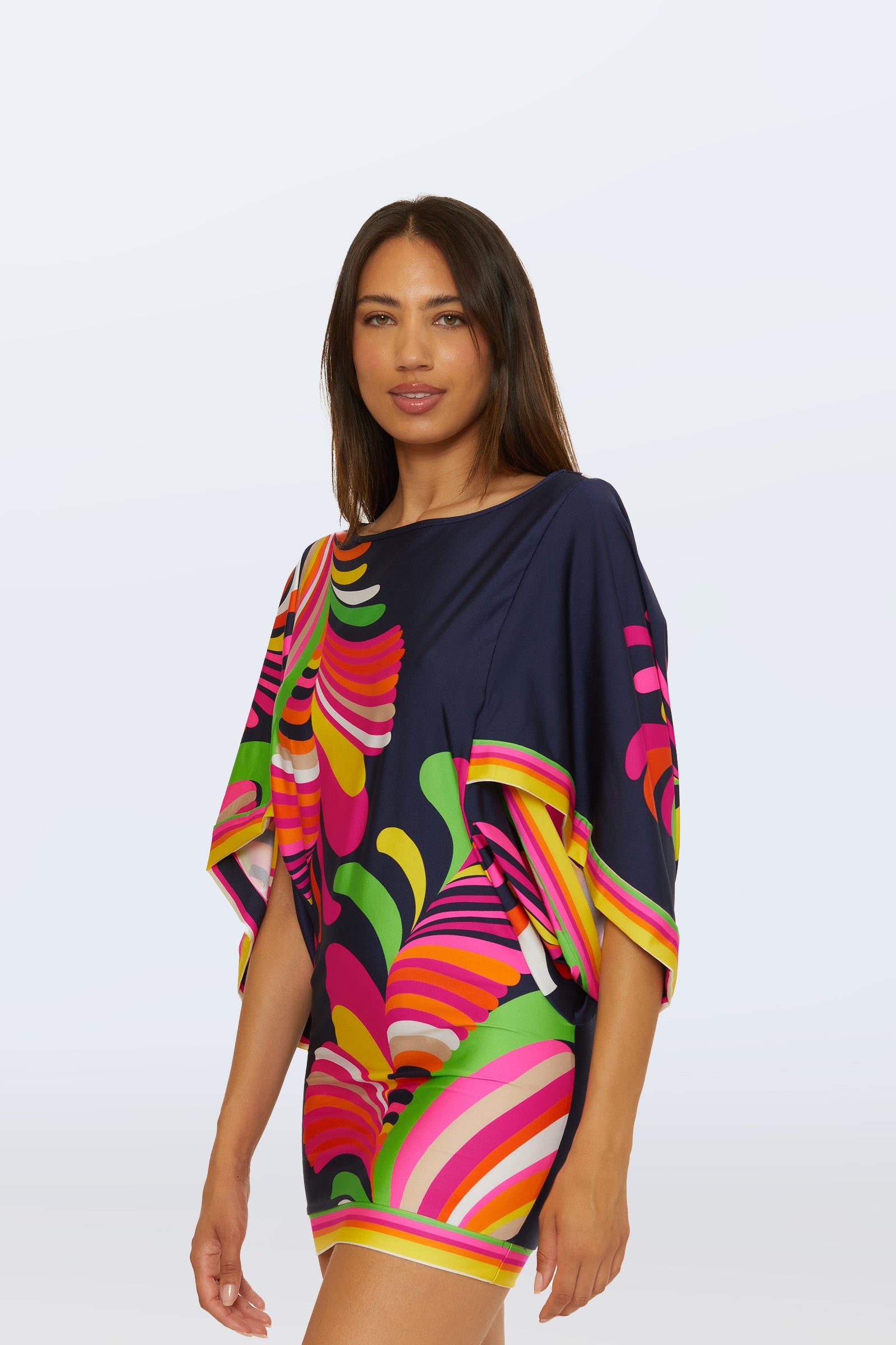 A woman with long brown hair models the Casablanca Swim Tunic in the Palms Paradise print, featuring a colorful abstract pattern of pink, yellow, green, and navy blue against a plain light background.