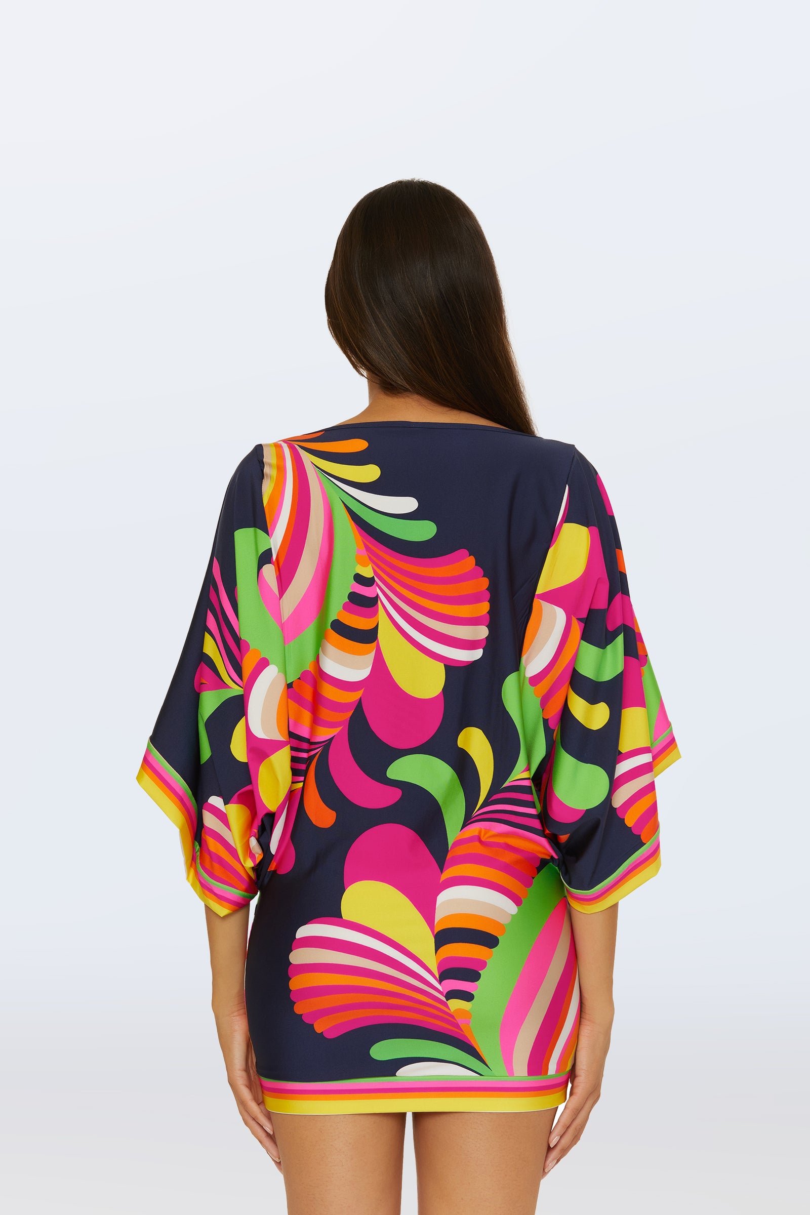 A woman with long dark hair faces away, wearing the Casablanca Swim Tunic - Palms Paradise, a short navy dress with wide sleeves and a bold abstract pattern in pink, yellow, green, and orange. The background is plain white.