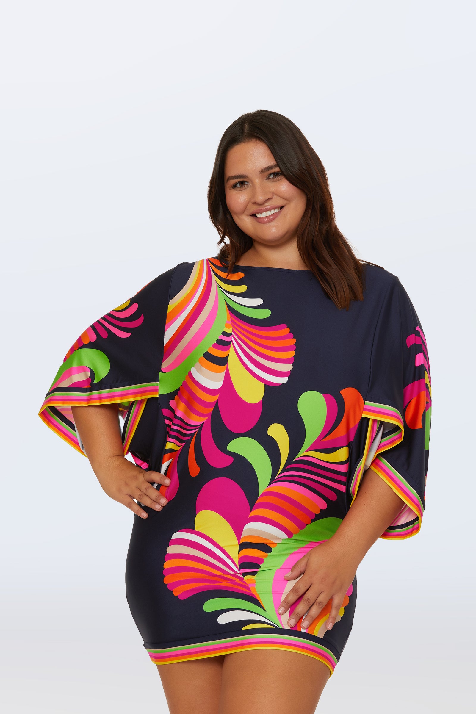 A smiling woman wears the Casablanca Swim Tunic - Palms Paradise, a short navy blue dress with bold, colorful wavy patterns, wide sleeves, neon trim, and stands confidently against a light background.