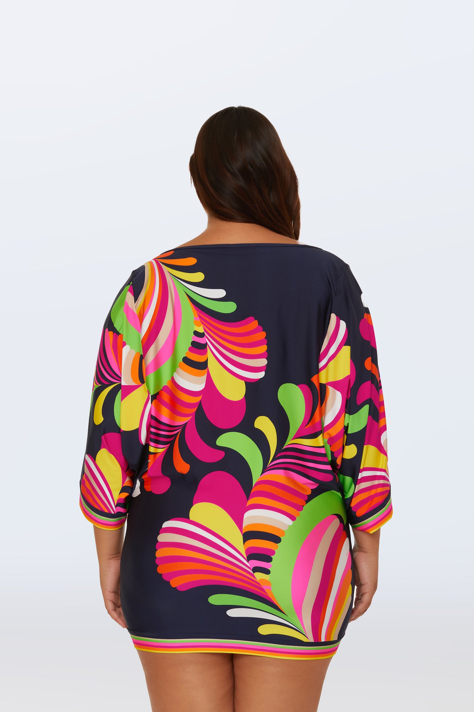 A person with long dark hair, seen from behind, wears the Casablanca Swim Tunic - Palms Paradise: a navy blue dress featuring bright, colorful abstract swirl patterns in pink, green, yellow, and orange on a plain light background.