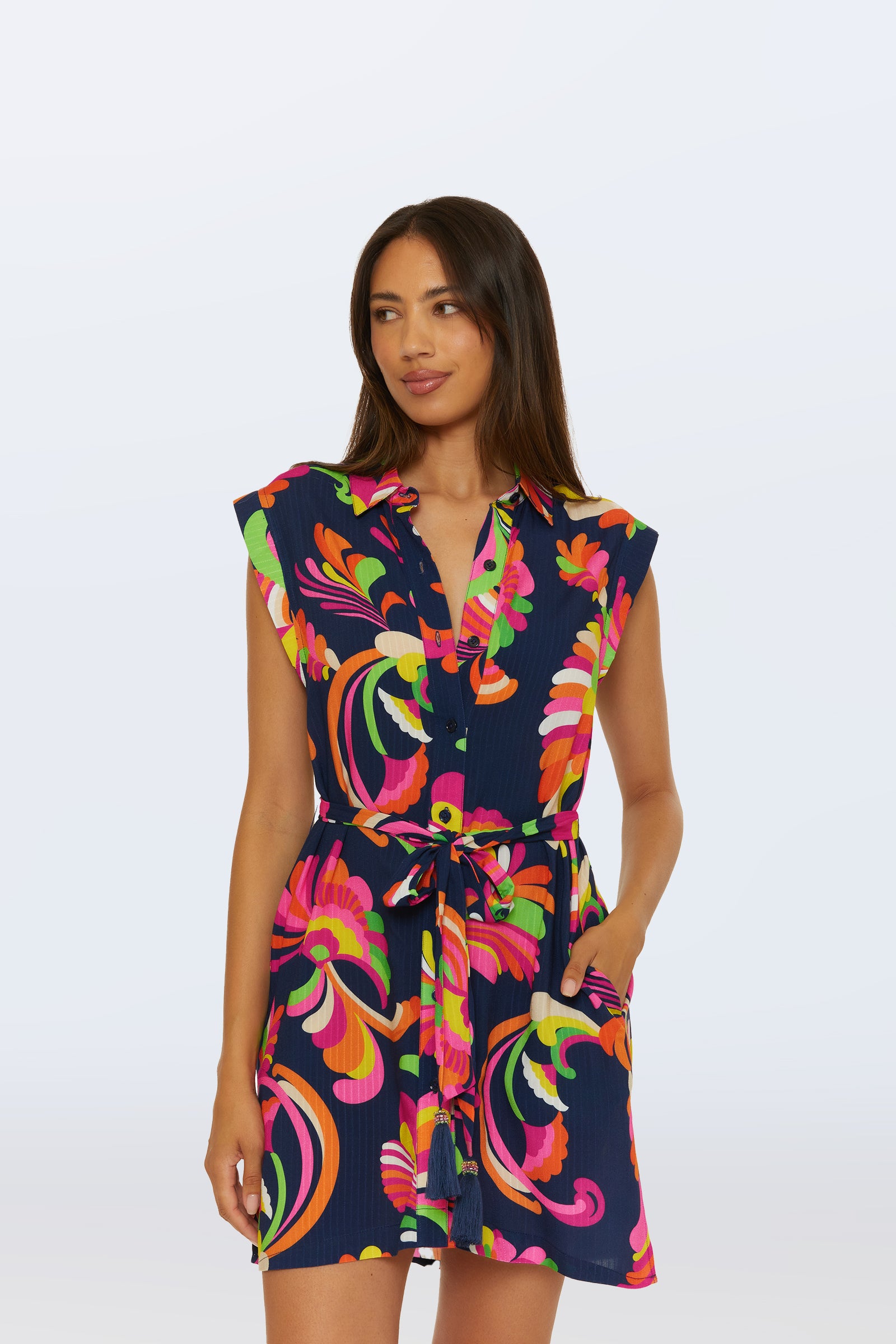 A woman wearing the Sleeveless Shirtdress Cover-Up in Palms Paradise—a multi-colored dress featuring a collared neckline and tie waist—stands against a plain light background.