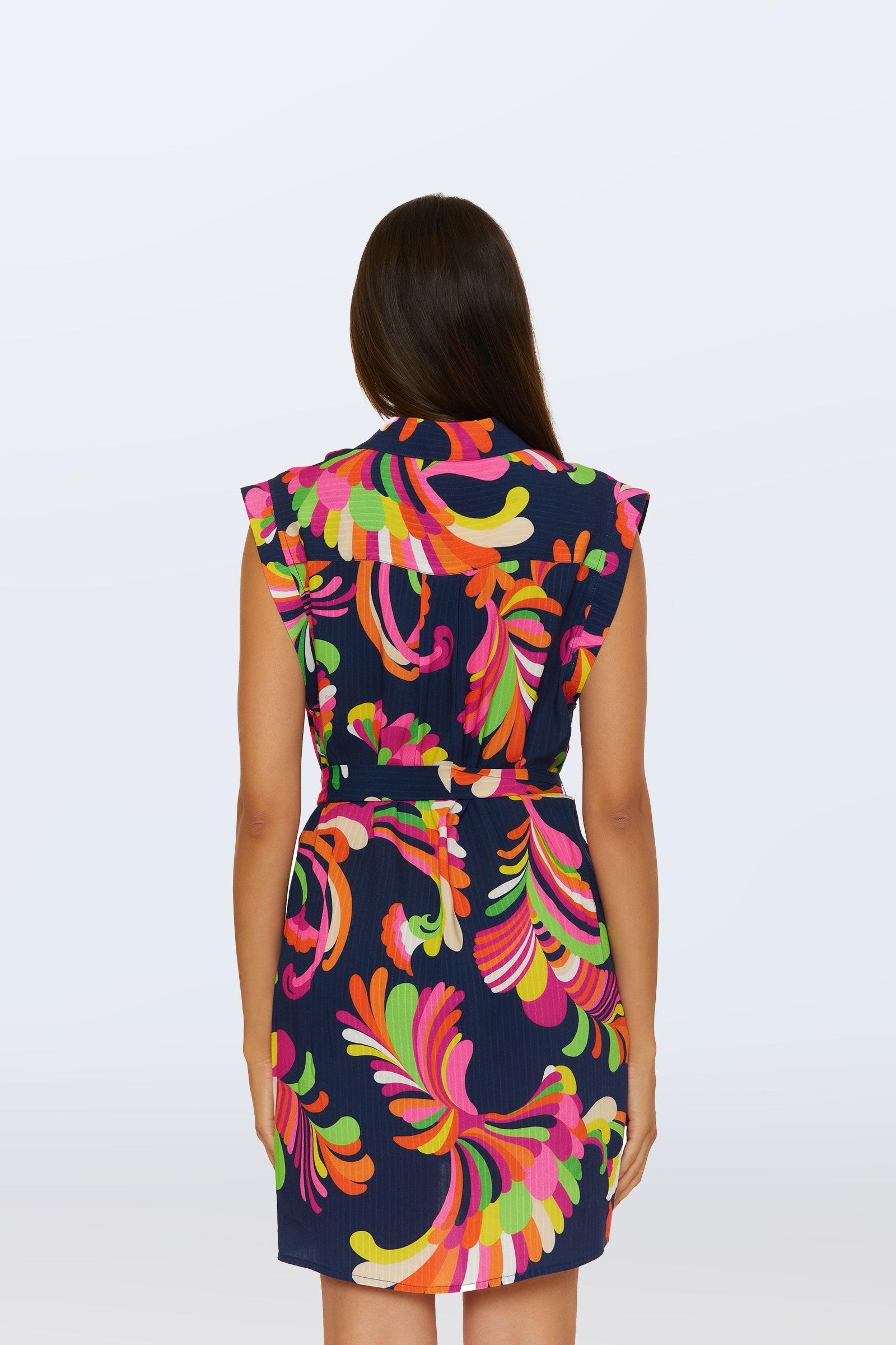 A woman faces away, wearing the Sleeveless Shirtdress Cover-Up in Palms Paradise—a navy dress featuring a vibrant abstract floral print in pink, orange, yellow, and green. It has a belted waist and the background is plain light gray.