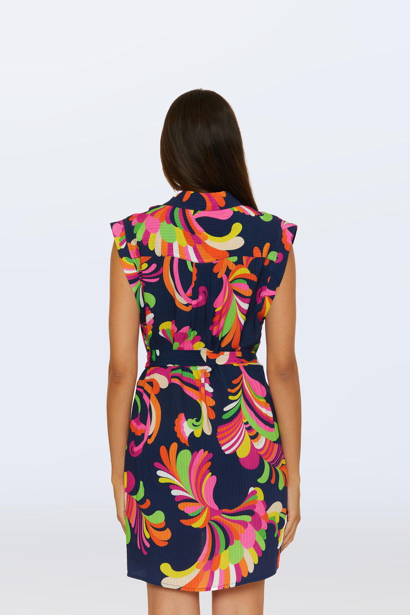 A woman faces away, wearing the Sleeveless Shirtdress Cover-Up in Palms Paradise—a navy dress featuring a vibrant abstract floral print in pink, orange, yellow, and green. It has a belted waist and the background is plain light gray.