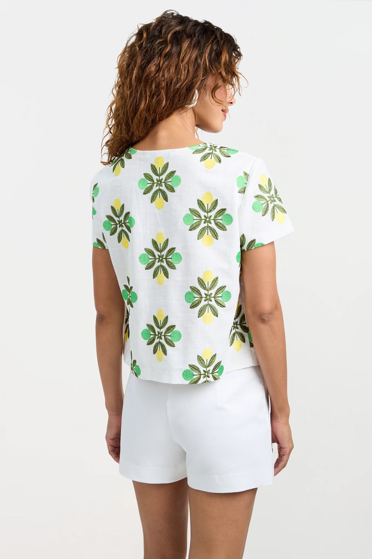 A woman with curly hair, seen from behind, wears the Salina Tie-Front Top—a white short-sleeve shirt with a green and yellow floral print—paired with white shorts against a plain light background.