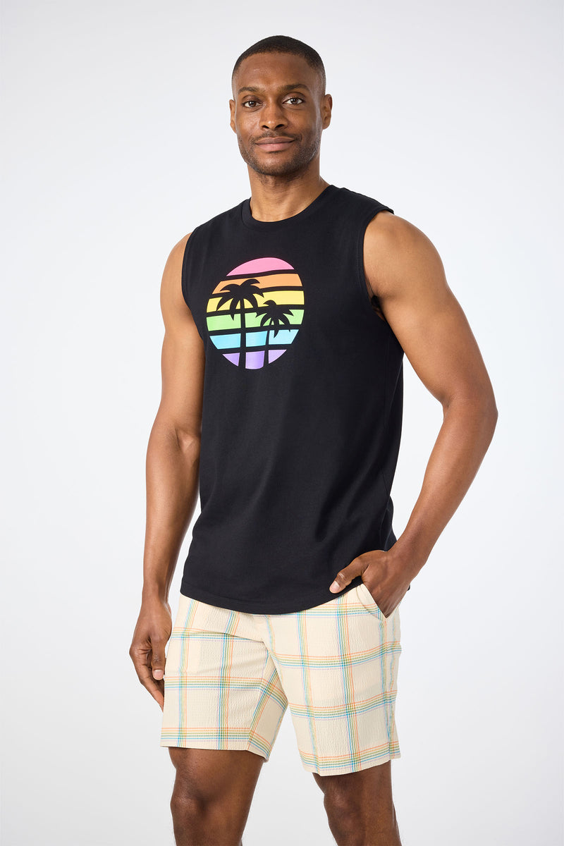 A man stands with one hand in his pocket, wearing the Sleeveless Muscle Tee - Pride featuring a rainbow-striped sunset and palm tree graphic, paired with light plaid shorts against a plain white background.