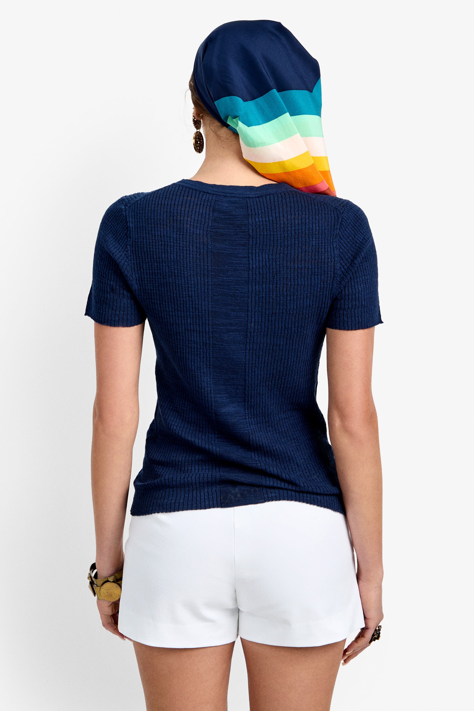 A person faces away, wearing the navy Mina Lace-Up Top with white shorts and a colorful striped headscarf. They accessorize with earrings and a bracelet against a plain white background.