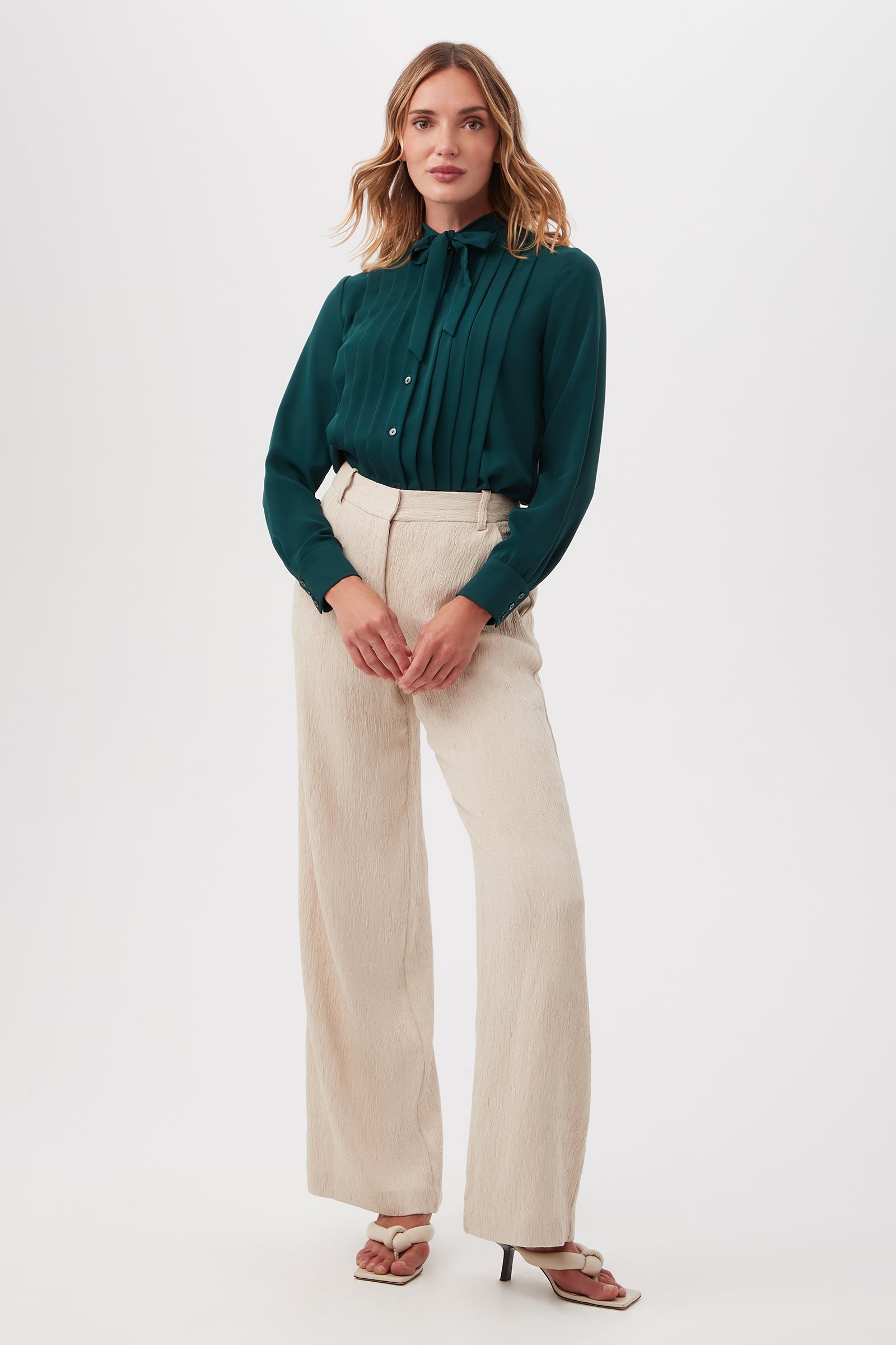 A woman stands against a plain white background wearing the Adaeh Top, a dark green blouse with pleated detail, styled with high-waisted cream wide-leg pants and open-toe heeled sandals. She poses with her hands clasped in front.