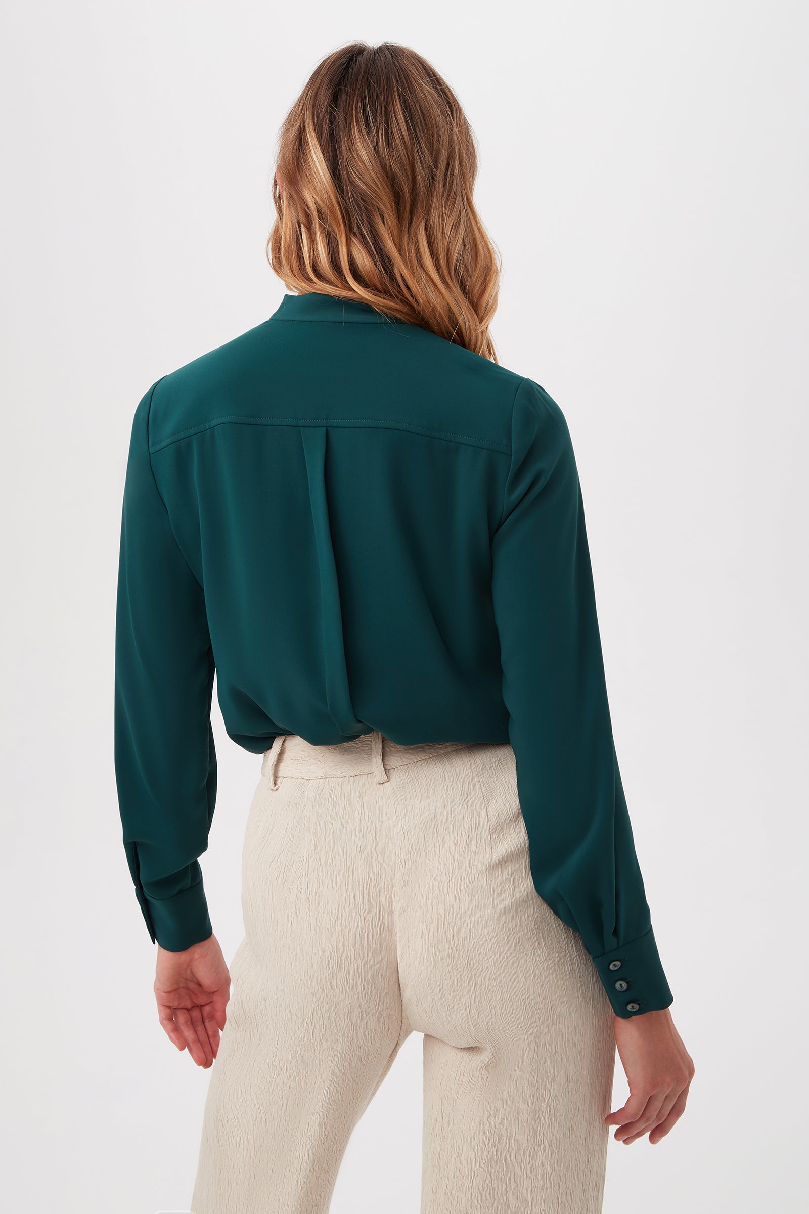 A woman with wavy, light brown hair is seen from behind, wearing the Adaeh Top in dark green tucked into high-waisted, light beige trousers against a plain white background.