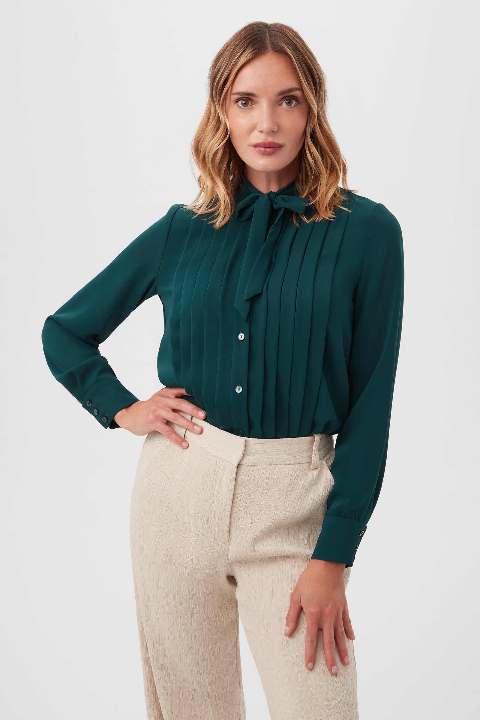 A woman with shoulder-length wavy hair wears the Adaeh Top in Victoria Lake, featuring long sleeves, a bow tie neck, and a pleated front. She pairs it with high-waisted, light beige textured pants and stands with one hand on her hip against a plain background.