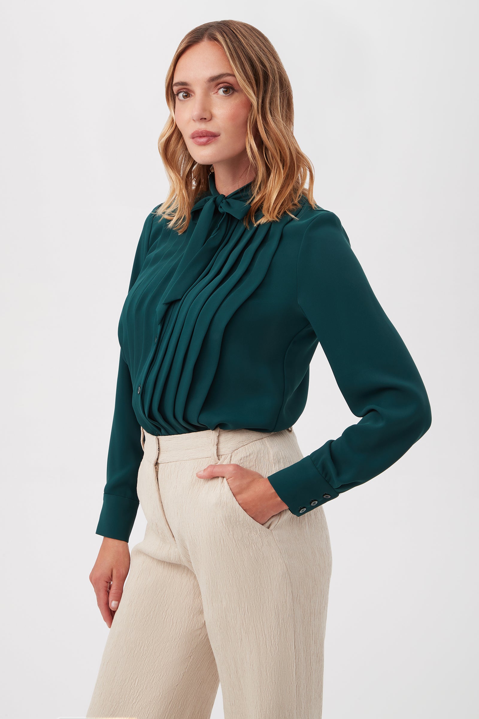 A woman with wavy, shoulder-length hair models the Adaeh Top—a dark green, long-sleeve blouse with pleats and a bow at the collar—paired with high-waisted, light beige trousers against a plain white background.
