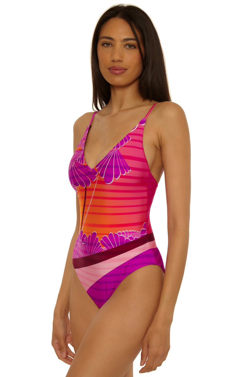A woman with long dark hair wears the Rhodes V Neck Maillot, a pink and orange one-piece swimsuit with floral and striped patterns, standing against a plain white background.