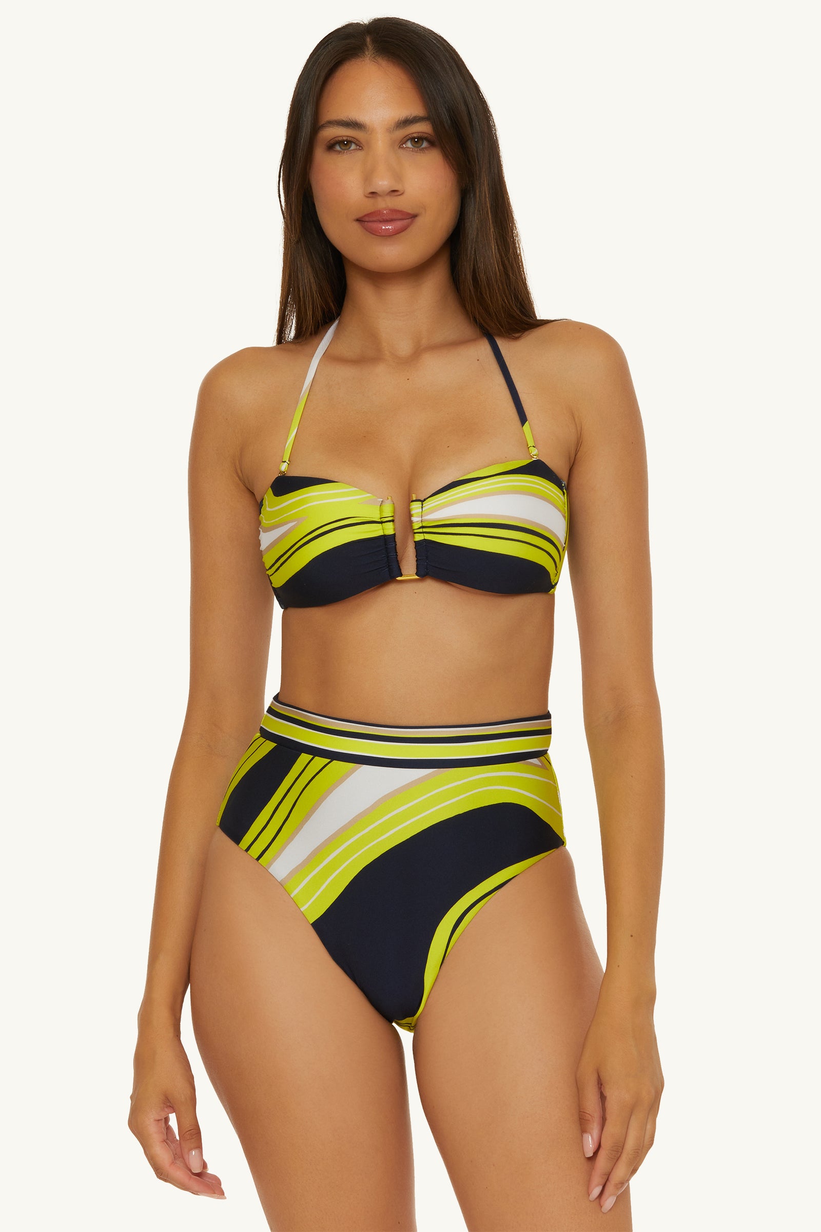 A woman models the Serena Swirl Strapless Bandeau Swim Top, featuring a bold yellow, navy, and white stripe pattern with a center cutout and high-waisted bottoms. She stands against a plain light background.