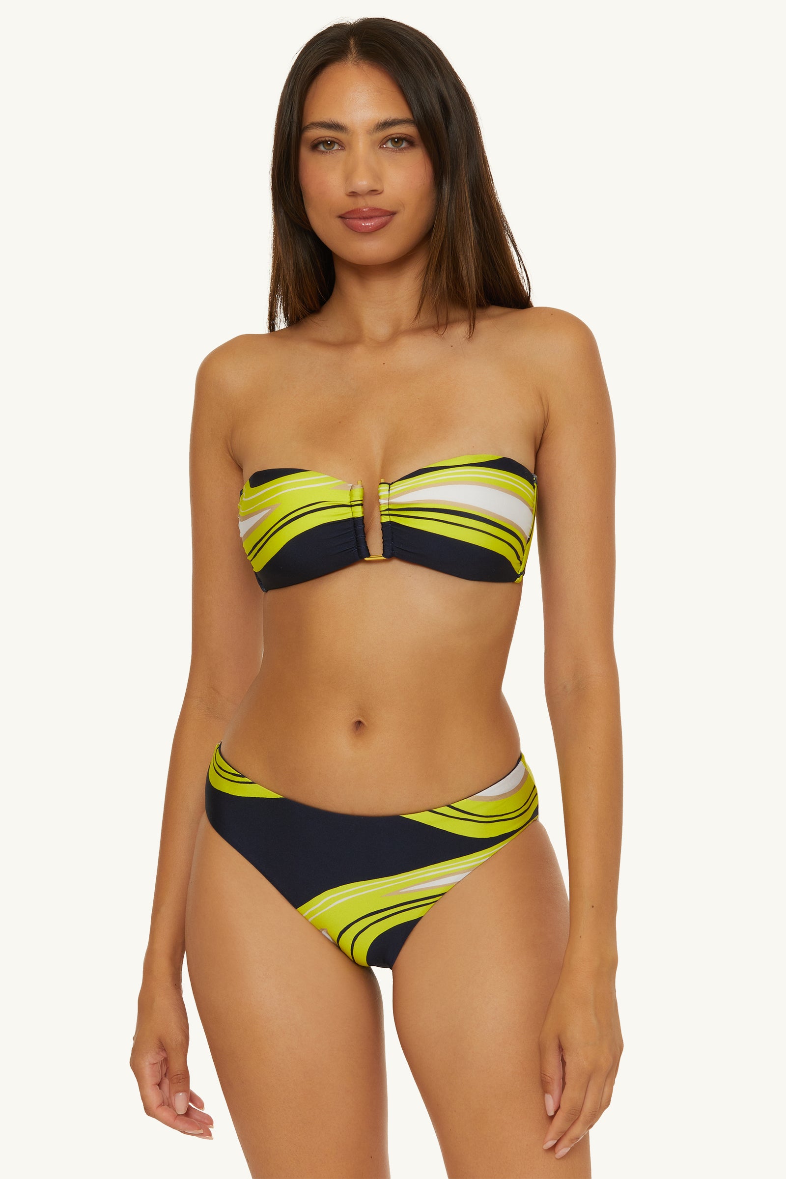 A woman with long straight hair and a neutral expression wears the Strapless Bandeau Swim Top - Serena Swirl, featuring a yellow, green, and black swirling pattern, center cut-out detail, and matching high-cut bottoms.