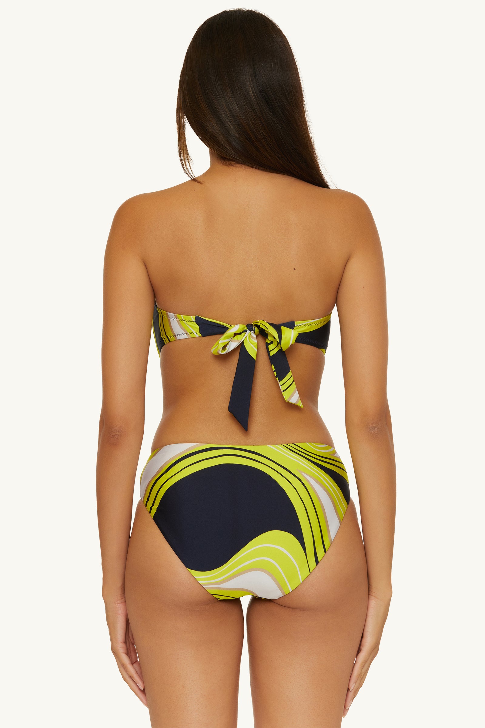 A woman with long brown hair is seen from the back wearing the Strapless Bandeau Swim Top - Serena Swirl, featuring a tie-back and a yellow, green, black, and white swirl pattern with high-waisted bottoms.