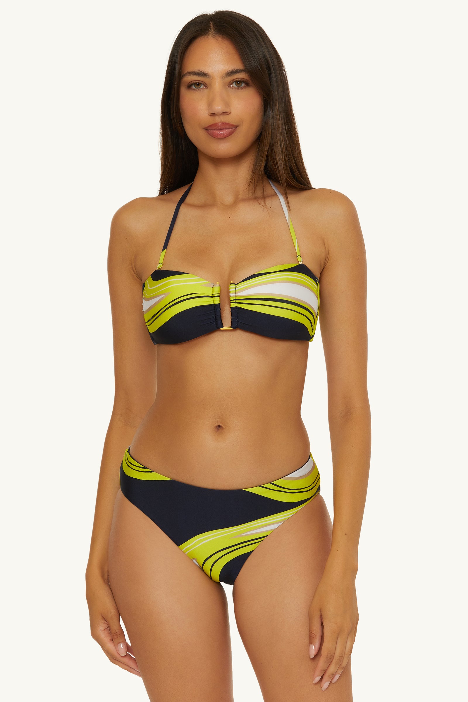 A woman with long brown hair and a neutral expression poses against a plain light background, wearing the Strapless Bandeau Swim Top in Serena Swirl Swim, featuring wavy green, yellow, black, and light gray patterns.