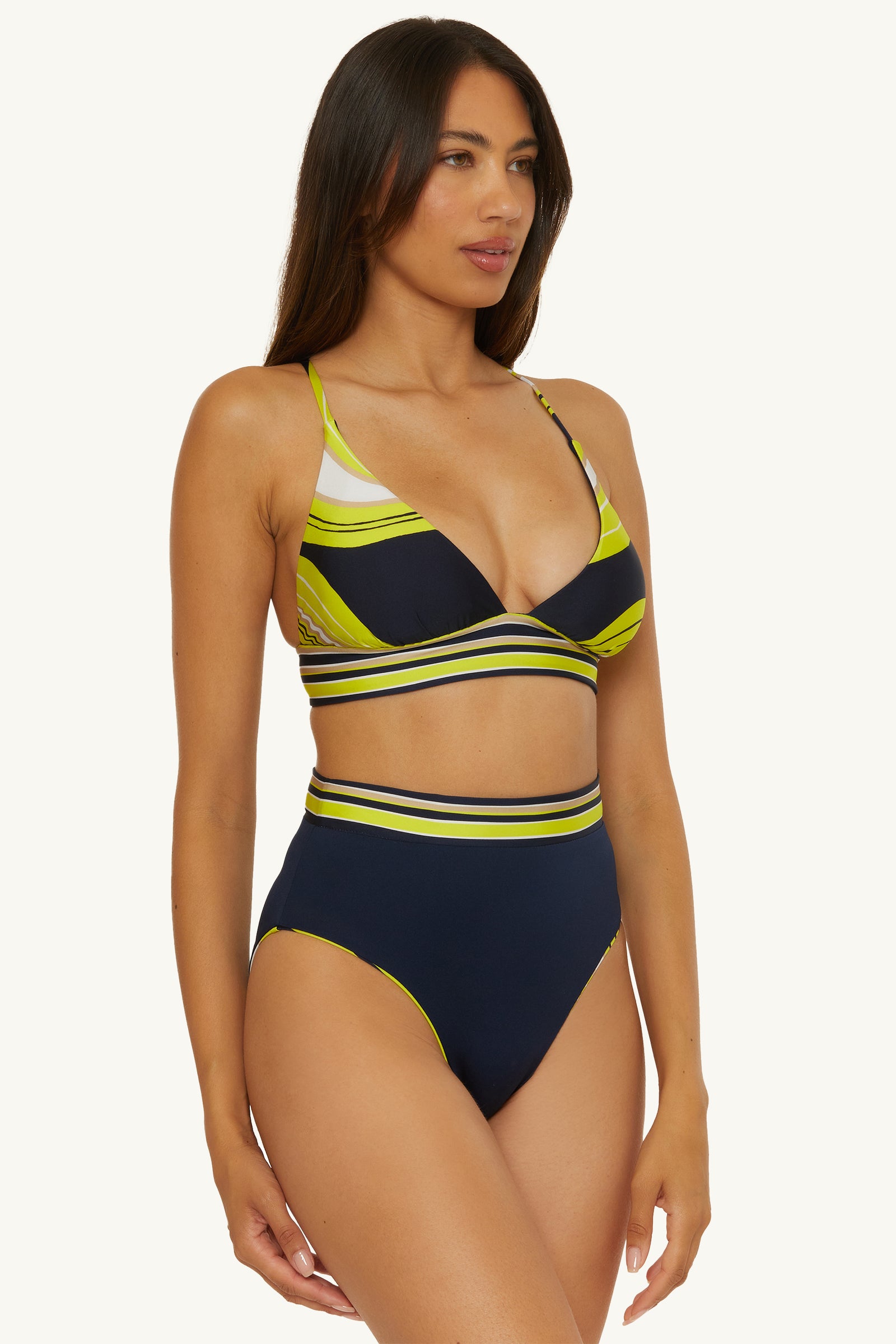 A woman with long brown hair models the Reversible High-Waist Swim Bottom - Serena Swirl, featuring a navy blue and yellow patterned bikini, against a plain light background.