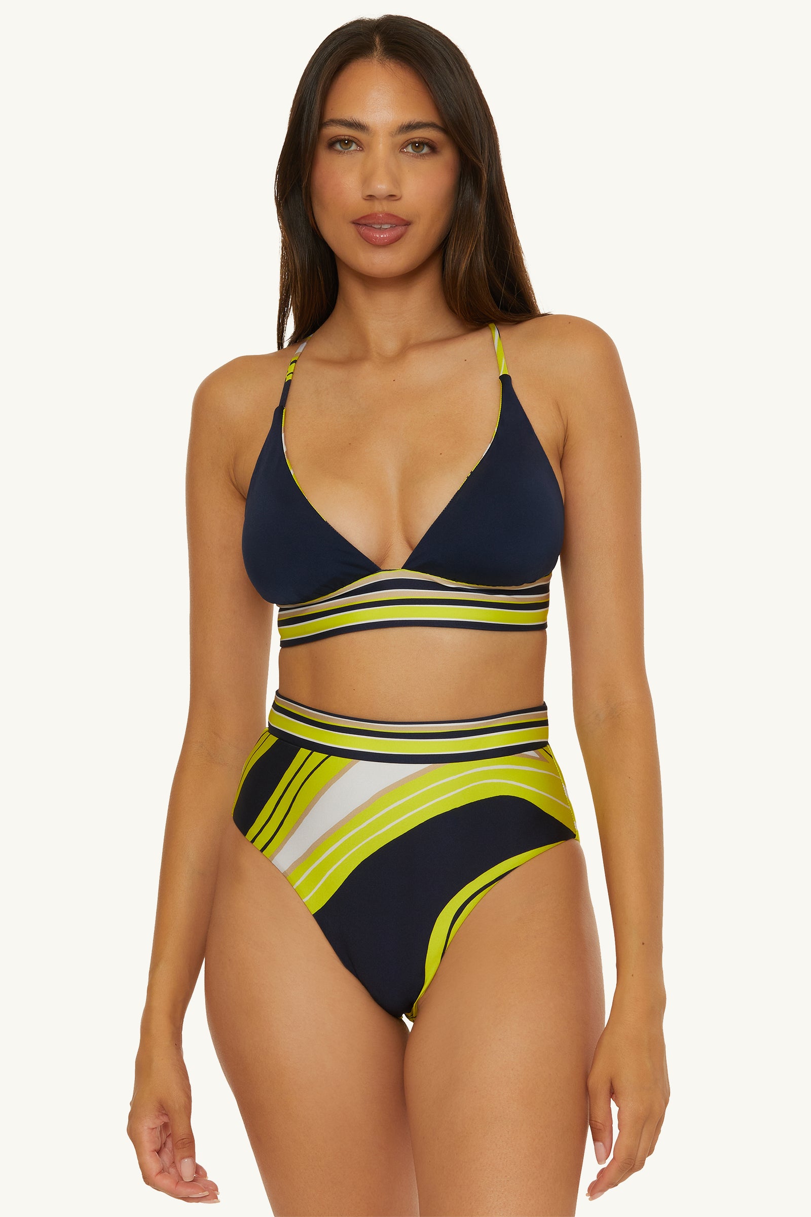 A woman wears the Serena Swirl Reversible Halter Swim Top, a navy bikini with yellow, white, and green geometric stripes and high-waisted bottoms, standing facing forward against a plain background.