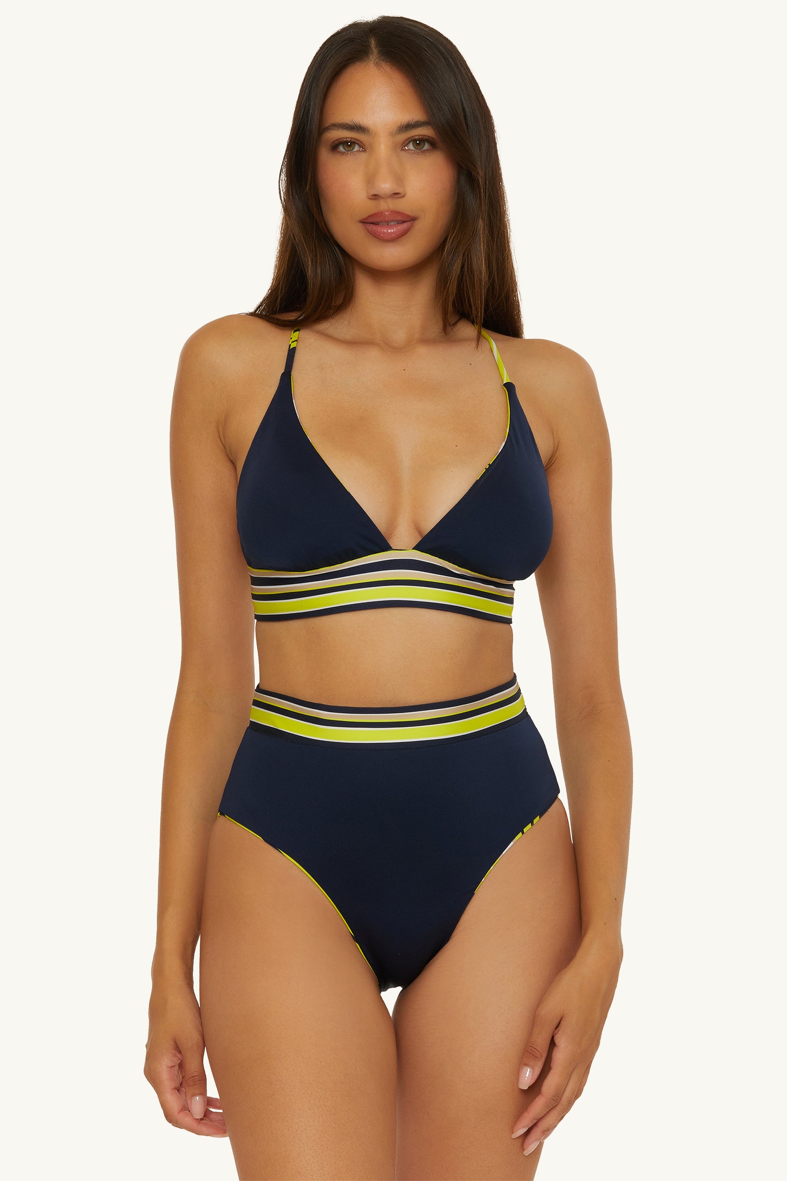 A woman with long brown hair models the Reversible Halter Swim Top - Serena Swirl, a navy blue bikini with yellow and white striped bands on the top and high-waisted bottom, standing against a plain light background.