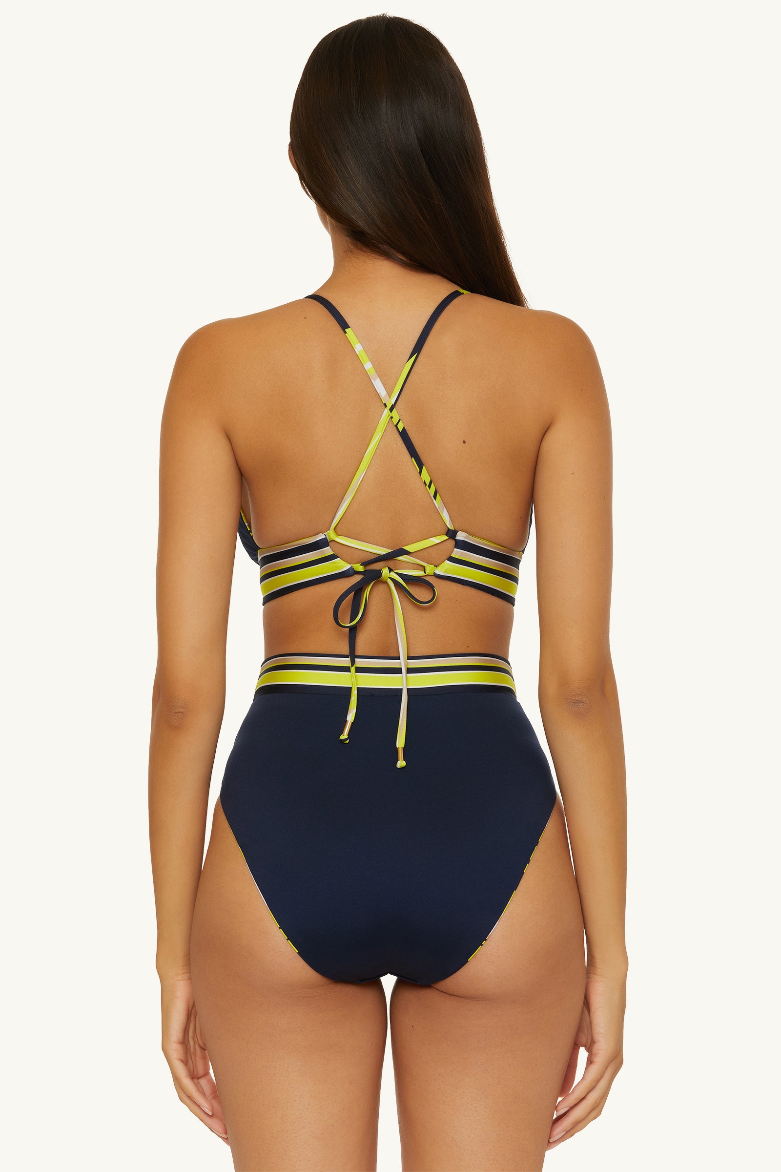 A woman with long brown hair is seen from behind wearing the Reversible High-Waist Swim Bottom - Serena Swirl, featuring navy blue fabric with yellow and green striped bands and crisscross straps that tie at the back.