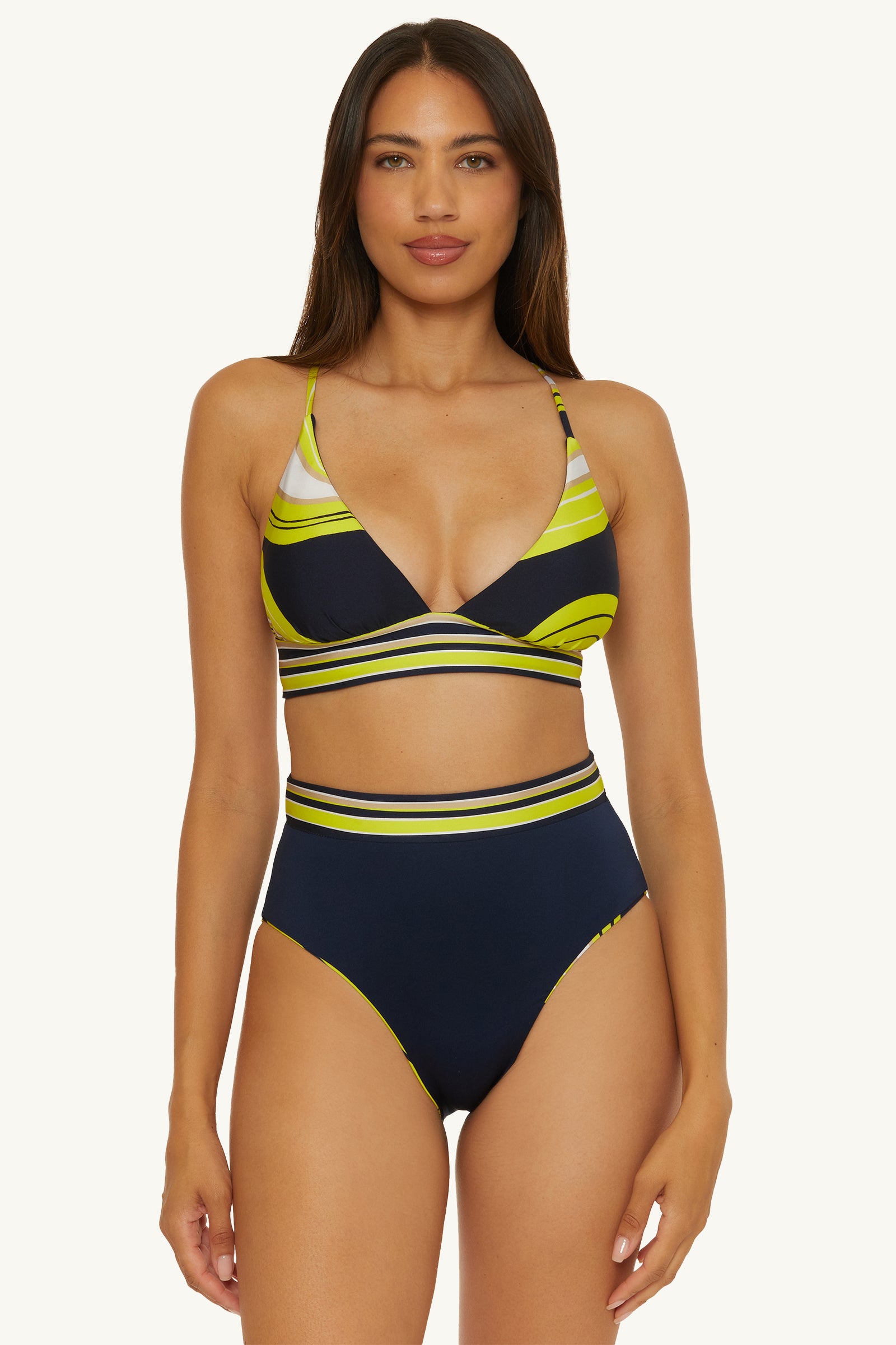 A woman faces forward against a plain light background, wearing the Serena Swirl Reversible High-Waist Swim Bottom paired with a navy blue and yellow striped, strappy v-neck bikini top.