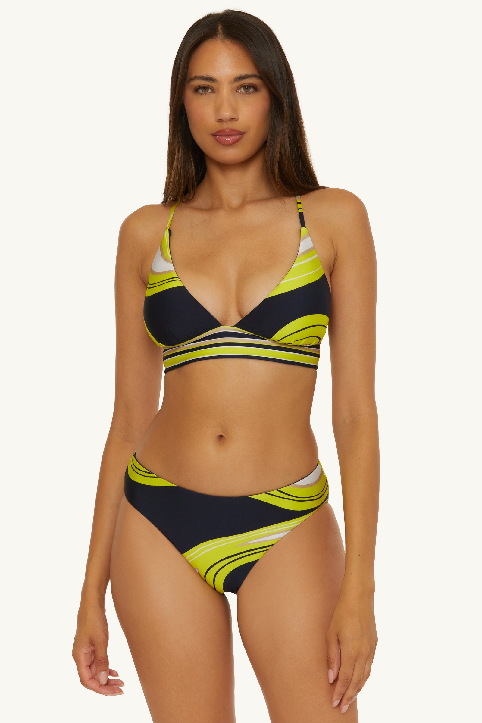 A woman with long brown hair and a neutral expression faces forward, wearing the Reversible Hipster Swim Bottom in Serena Swirl—a black bikini with green and yellow abstract swirls, featuring high-waisted bottoms and a triangle top with straps.