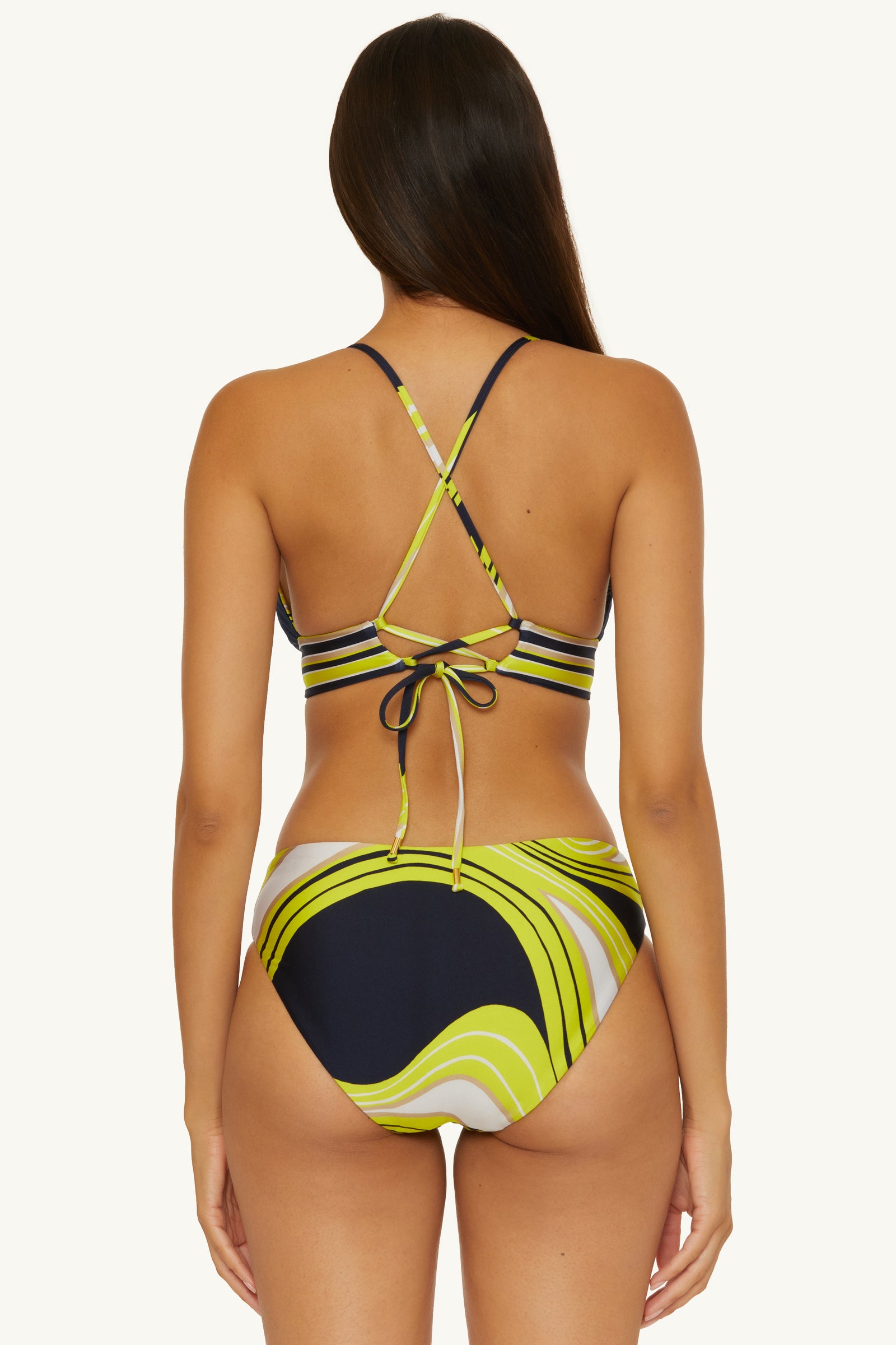 A woman with long brown hair is seen from behind wearing the Serena Swirl Reversible Hipster Swim Bottom, featuring a retro black, lime green, and white wavy pattern. The bikini top has crisscross straps and ties at the back.